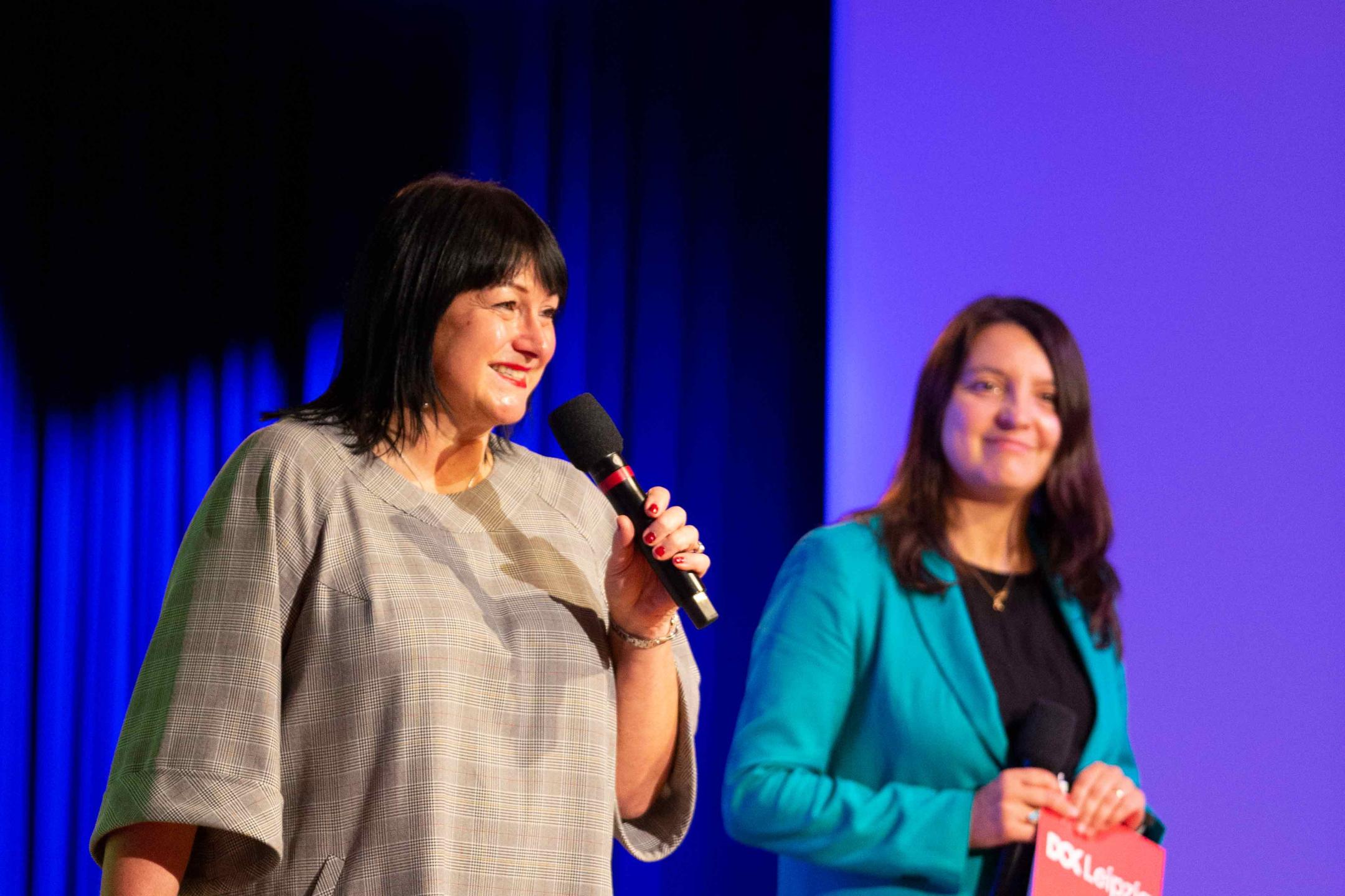 Two women are presenting on a stage with a background in blue. Both are smiling, the woman at the left is holding a microphone.