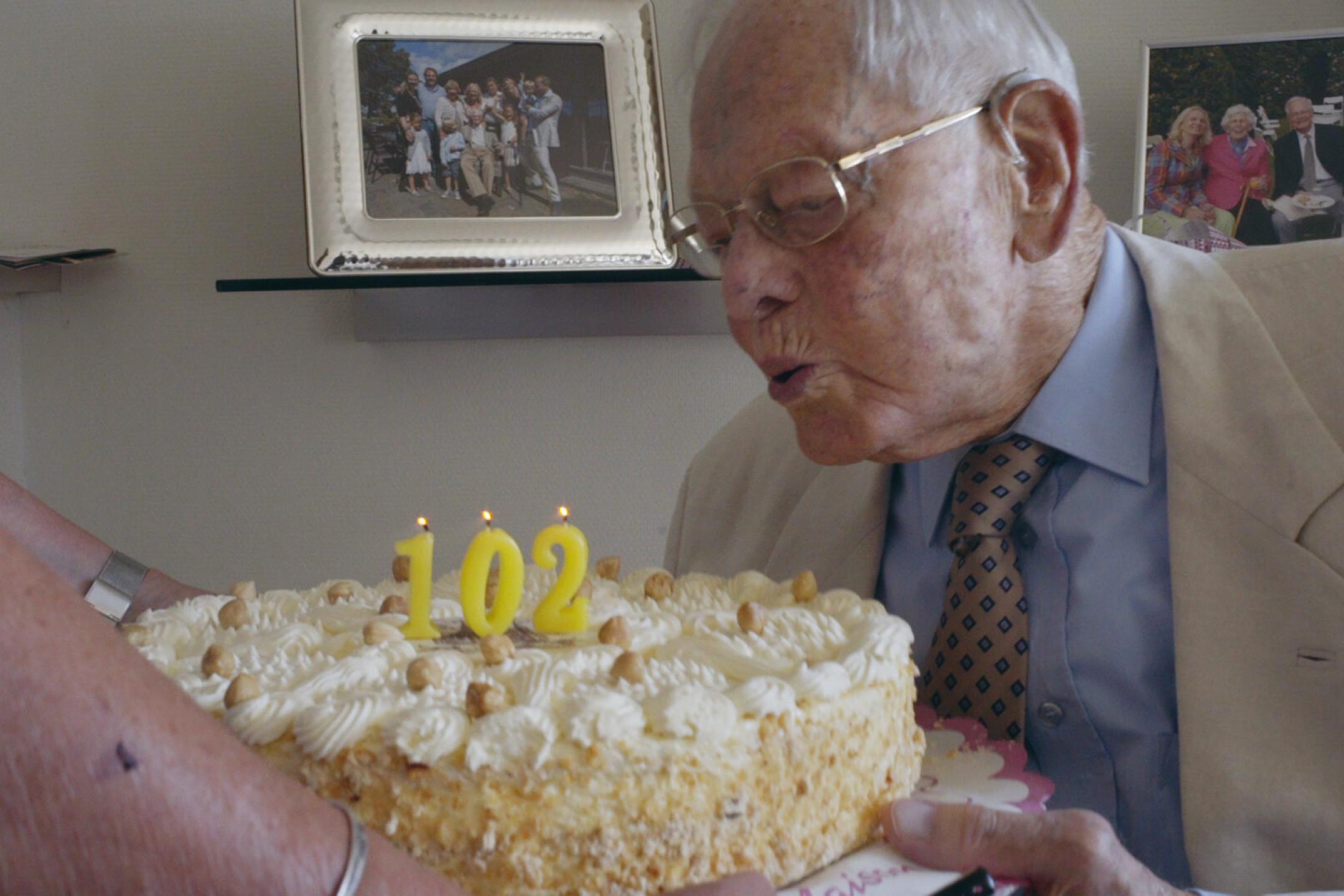 An older man blows out the candles on a huge birthday cake made of cream. There are three candles with the numbers 1, 0 and 2.