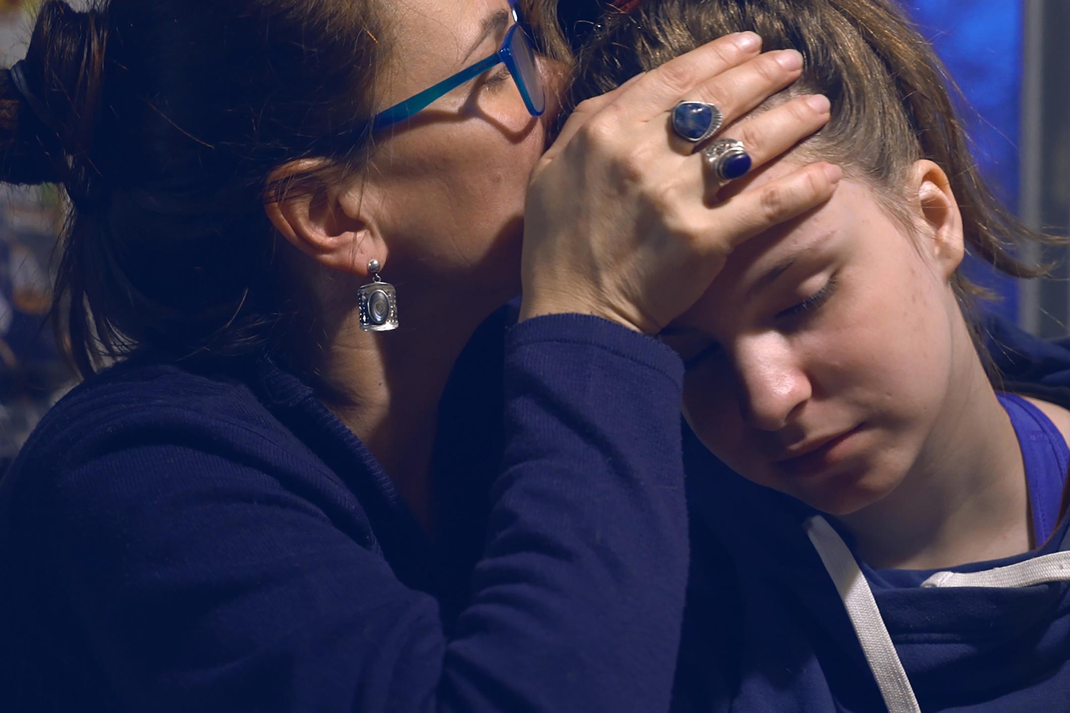 A woman holds a teenager girl in her arms and kisses her forehead. The teenager has her eyes closed.