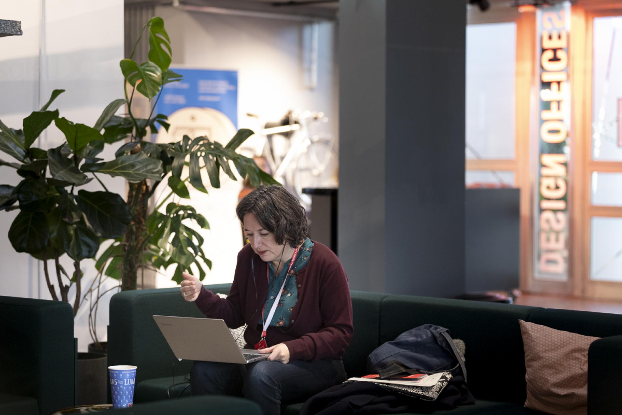 A woman with earpods sits on a couch with a laptop on her knees. She leans a bit forward and looks at the laptop display.