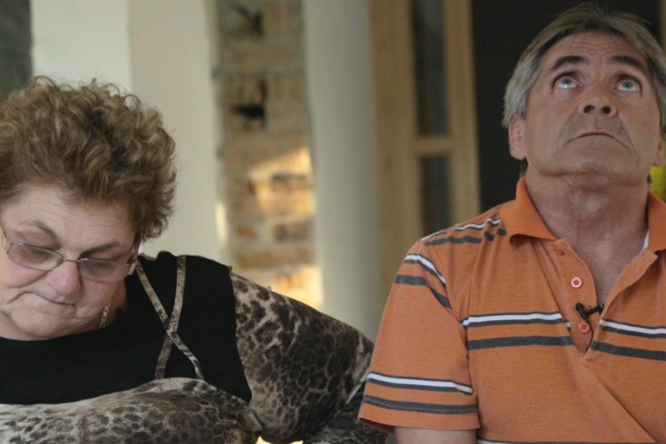 An elderly couple is sitting next to each other. The woman (left) is looking down, the man is rising his head and eyes up to the sky.