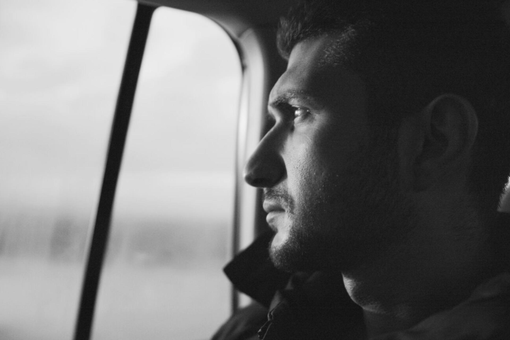 A black-and-white pic: The close-up of a man’s face in profile. He is looking out of a car window.