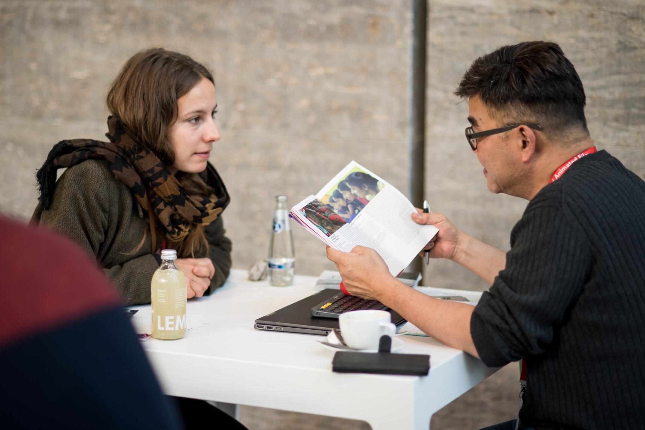 A woman and a man are sitting opposite each other at a small white working table. He is looking into a brochure with photos, she is slightly leaning forward looking at him.