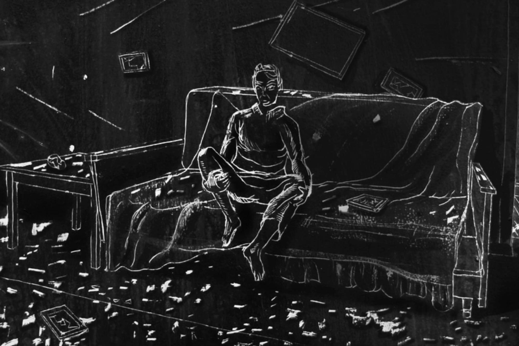 A room in black-and-white where a person sits on a couch that is covered with a blanket.