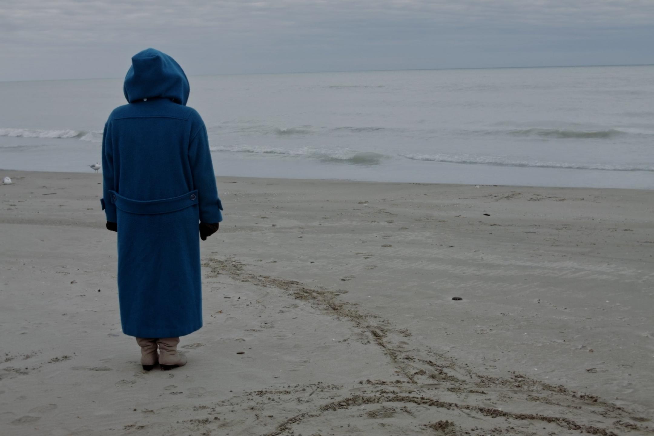 Am Strand, vor dem flachen, ruhigen Meer, steht eine Person in beinahe bodenlangem blauen Regenmantel mit dem Rücken zur Kamera. Sie hat die riesige Kapuze hochgezogen und schaut zum Meer.