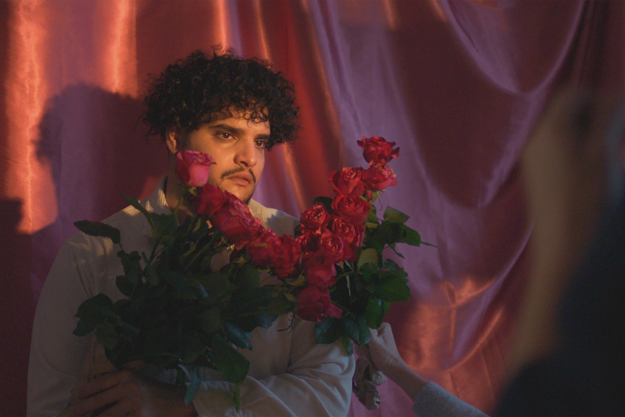 Ein Mann mit dunklen Locken steht vor einem rosa schillernden Vorhang und hält fünf Rosen vor sich. Eine andere Hand hält weitere Rosen hoch, sodass sie ein V um sein Gesicht bilden.