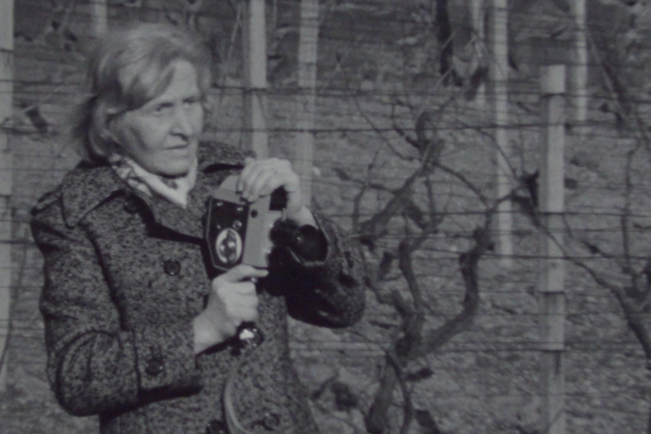 Schwarz-weiß-Aufnahme: Eine ältere Frau in einem Wintermantel hält eine Super-8-Kamera in der Hand und schaut in die Ferne.