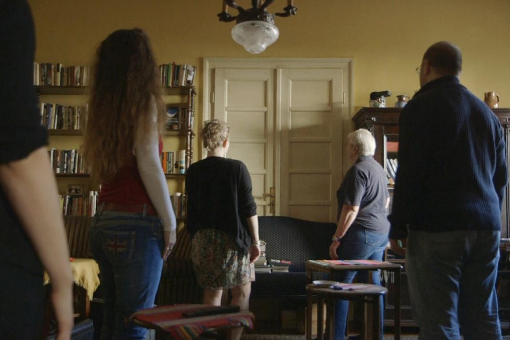In einem gelb gestrichenen Wohnzimmer stehen 5 Menschen unterschiedlicher Generationen weggedreht von der Kamera und schauen auf eine geschlossene Flügeltür