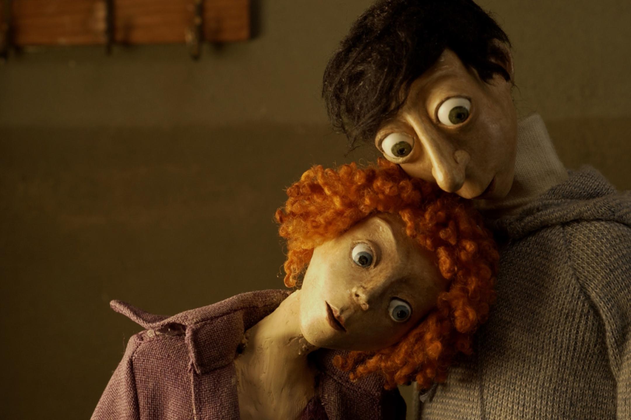 Ein Still aus einem Puppentrickfilm. Zwei Puppen stehen nebeneinander, die weibliche mit roten Locken hat ihren Kopf an die Brust der männlichen Puppe gelehnt. Beide haben sehr große Augäpfel, die hervorquellend auf etwas außerhalb des Bilden schauen