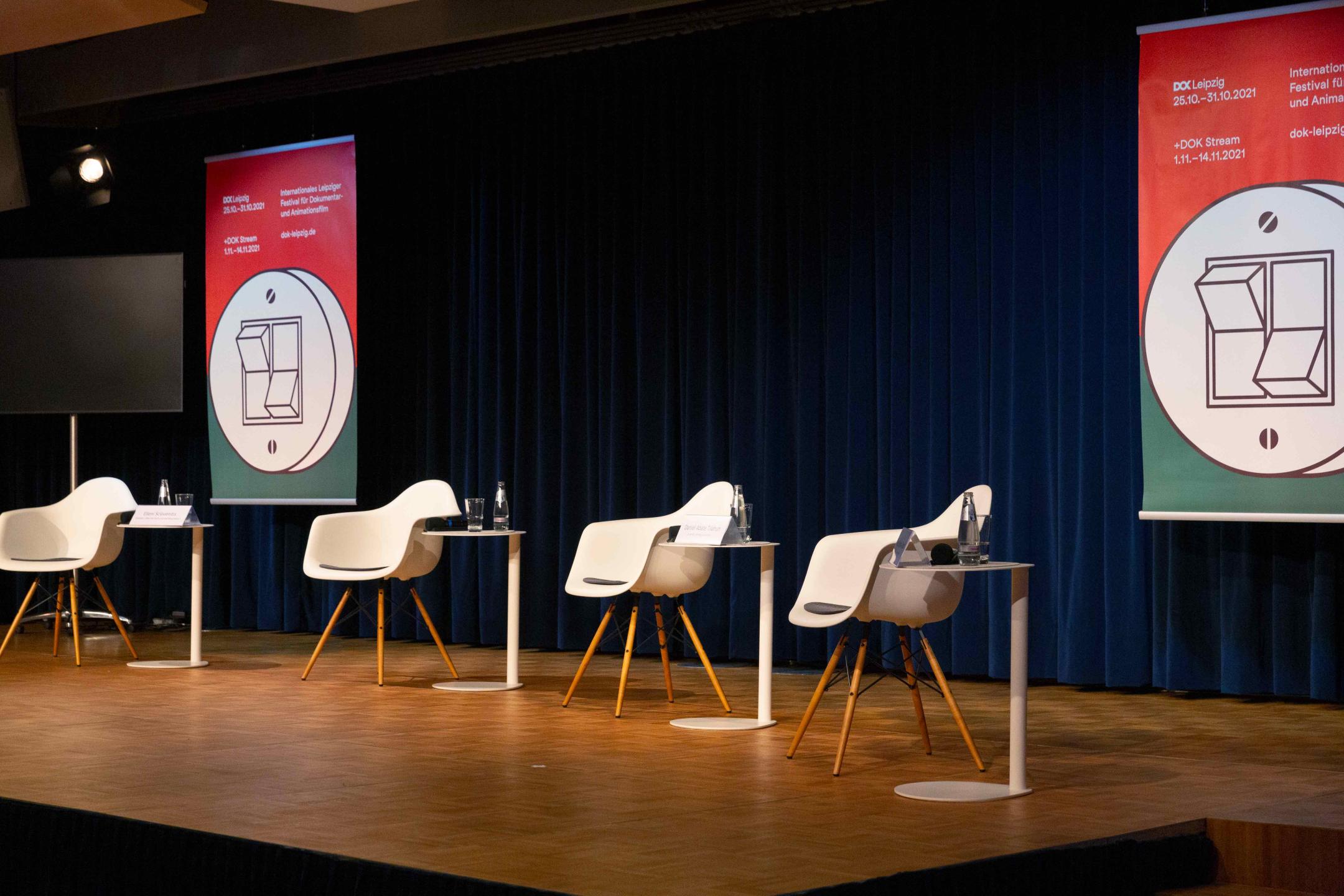 A podium prepared for a panel: Four empty white chairs with small tables in between and a DOK Leipzig festival poster in the background .