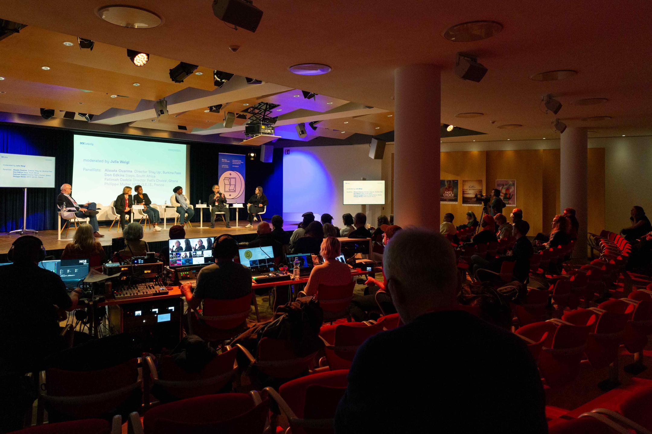 An event room. Looking across the rows of seats toward the podium, where five people are sitting with handheld microphones, small white side tables between them. The rows of seats are well filled. At the front of the first row, a large live editing station with various monitors.