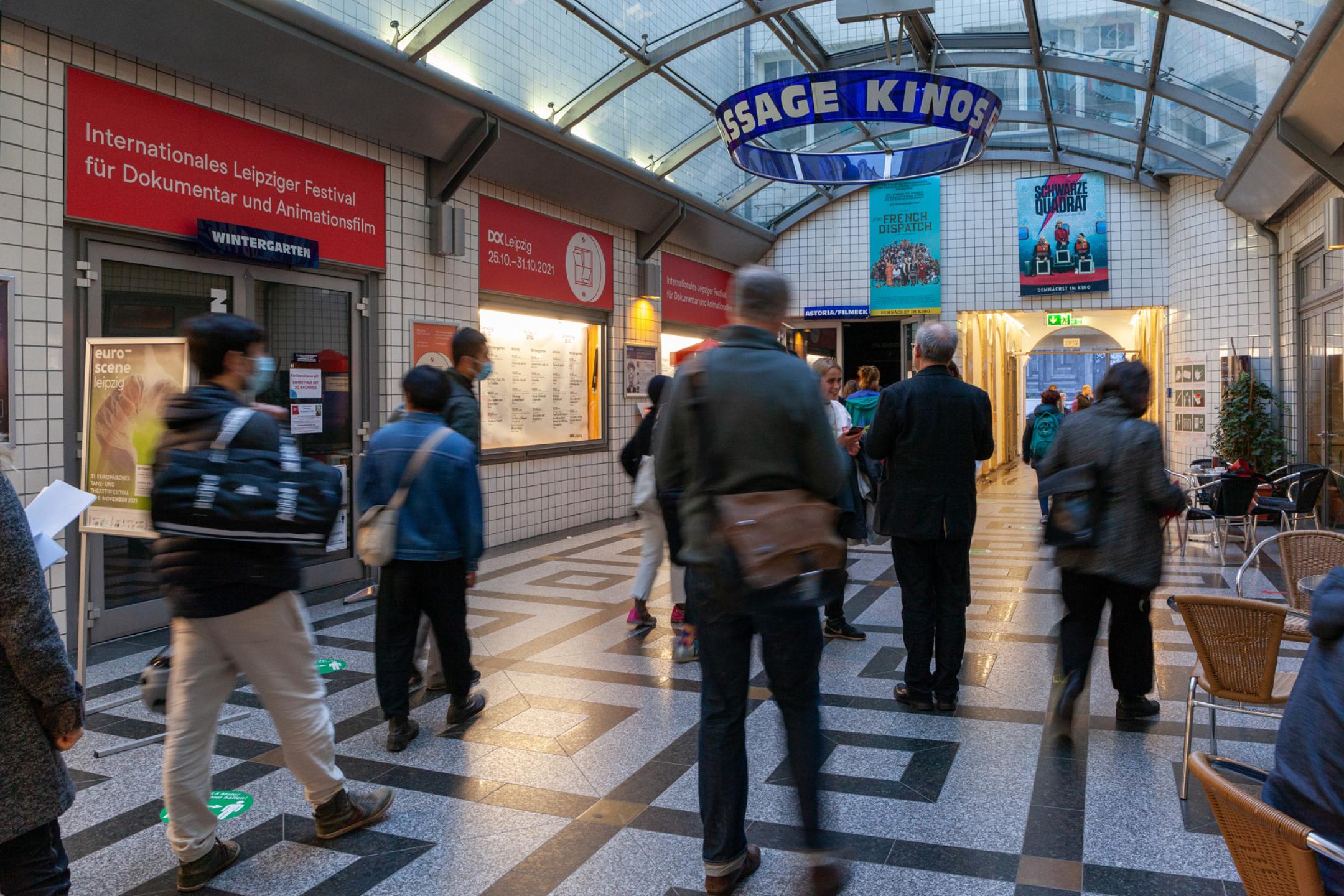 Mehrere Personen gehen durch einen glasüberdachten breiten Gang in einer Passage. Oben hängt ein blauer Ring mit der Aufschrift "Passage Kinos". Links ein rotes Banner mit der Schrift "Internationales Leipziger Festival für Dokumentar- und Animationsfilm".