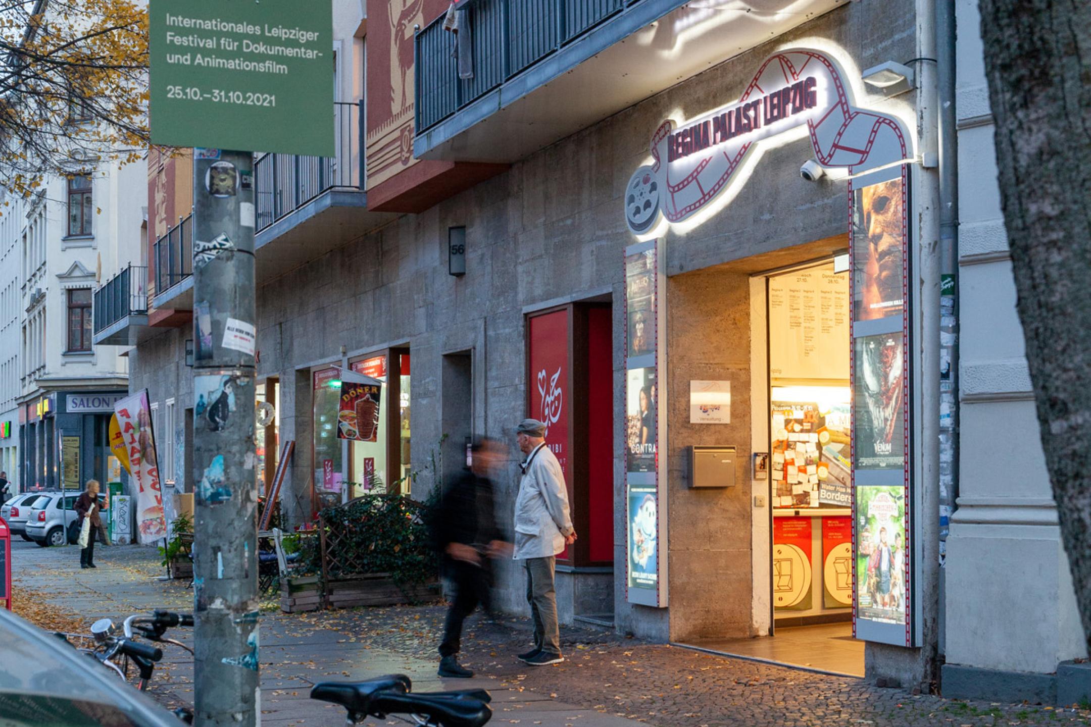 Ein beleuchteter Eingang zu einem mehrstöckigen Haus. Am Eingang sind mehrere Schaukästen, darüber steht "Regina Palast Leipzig".