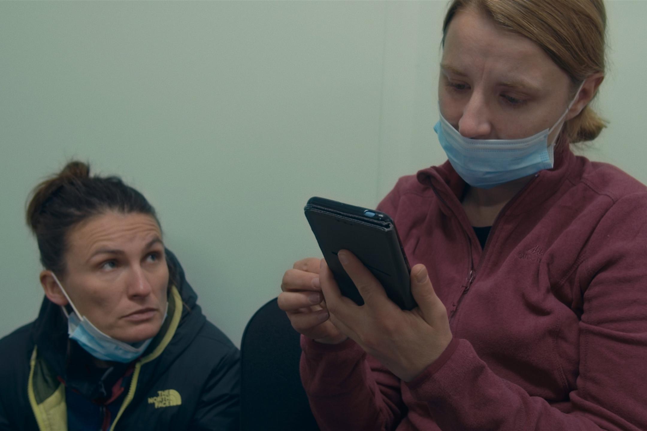 Eine Frau mit Nase-Mund-Schutz schaut auf ihr Handy, neben ihr sitzt eine weitere Frau, die gebannt in ihr Gesicht schaut.