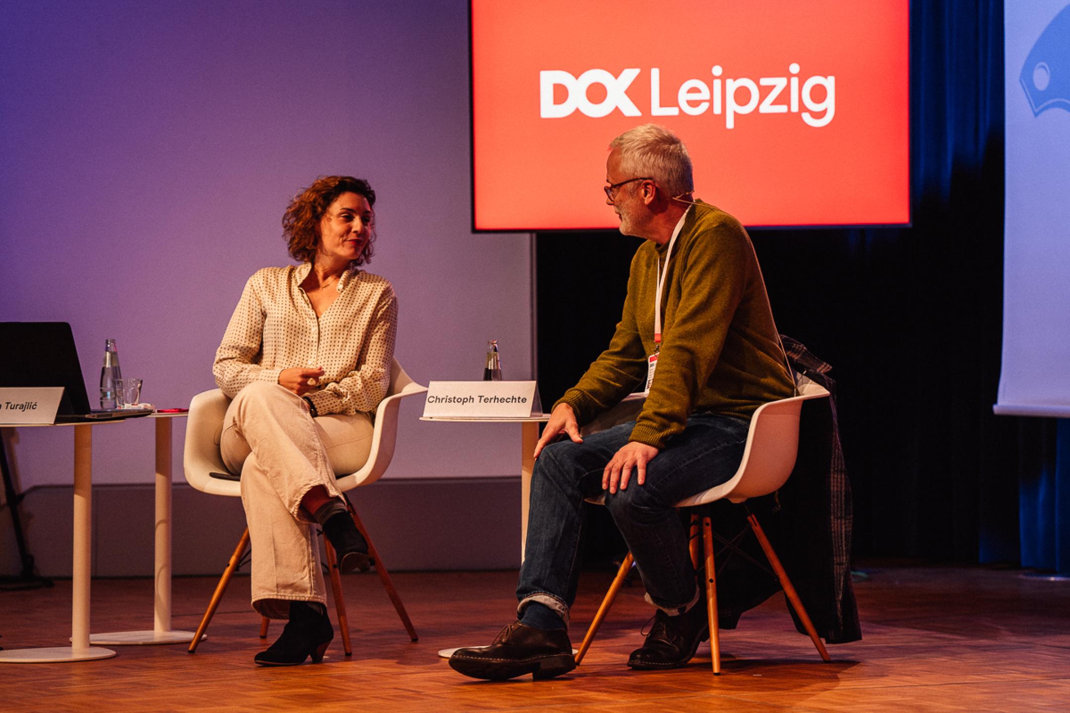 Eine Frau und ein Mann sitzen nebeneinander auf einem Podium. Sie haben sich einander zugewandt und sprechen mit einander. Im Hintergrund ist ein Bildschirm mit dem Logo von DOK Leipzig.