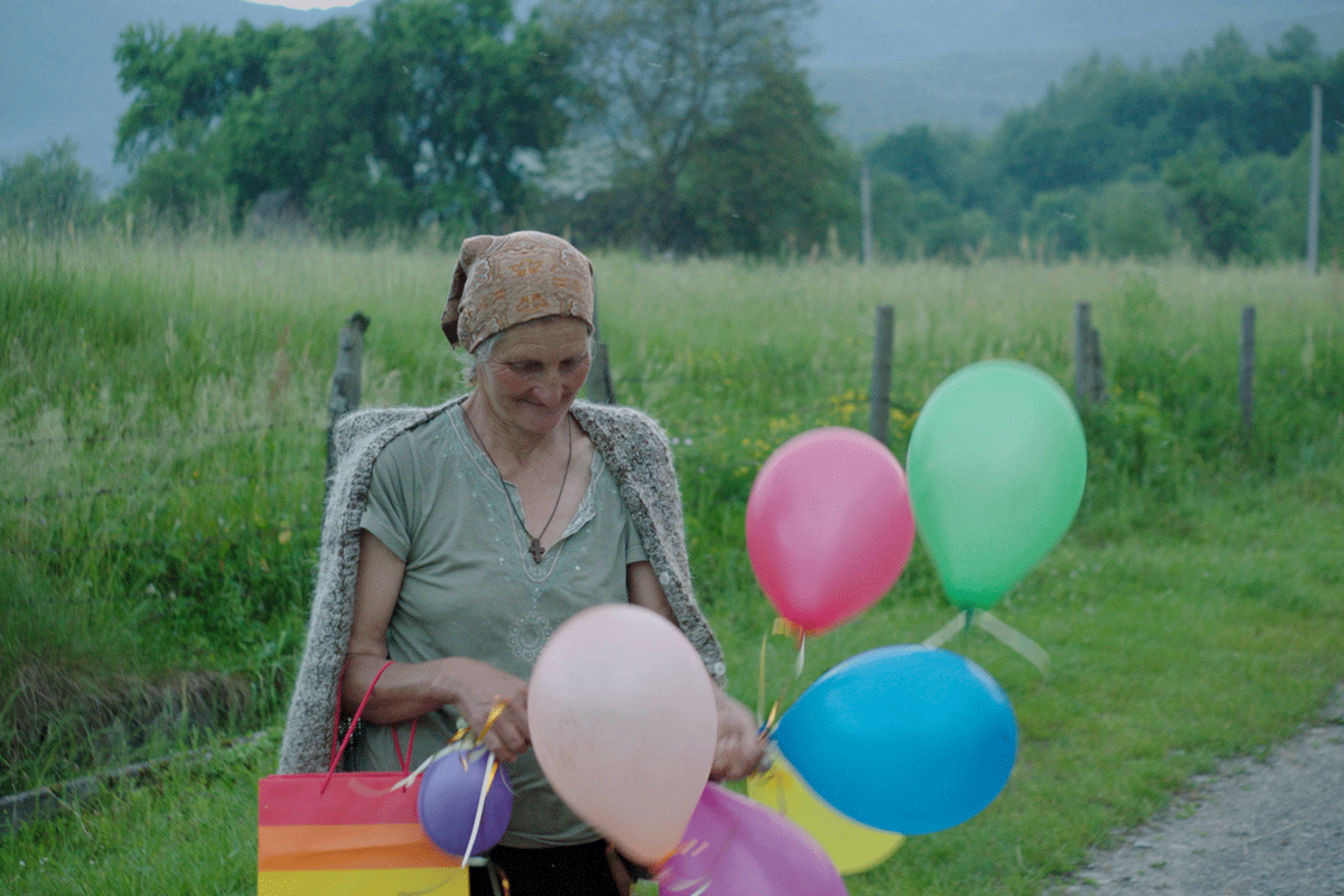 Eine ältere Frau steht vor einem Feld und hat ein Bündel an bunten Luftballons in der Hand. die um sie herumschweben.