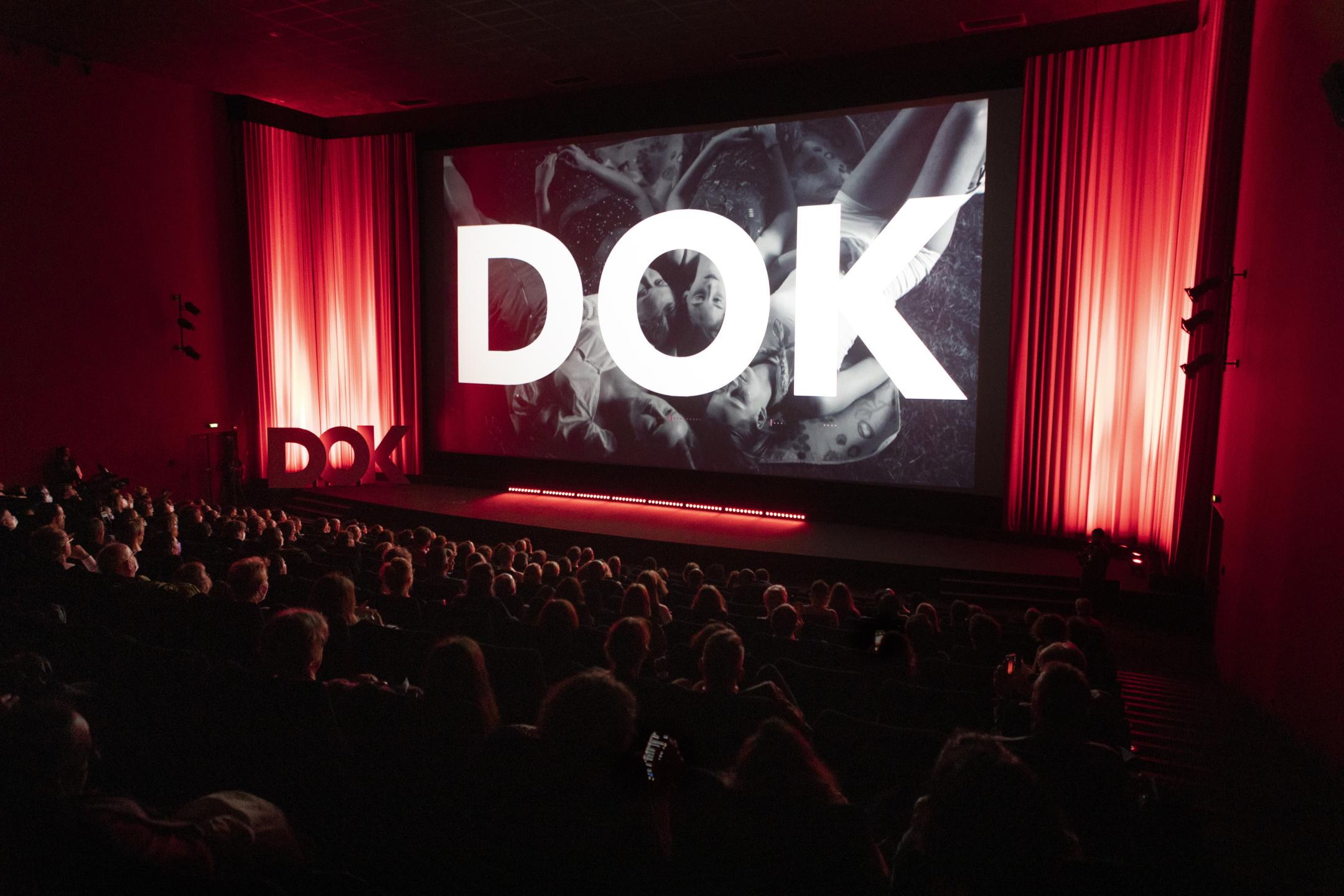In einem Kinosaal mit vollbesetzten Reihen ist auf der Leinwand riesengroß das Wort Dok zu sehen