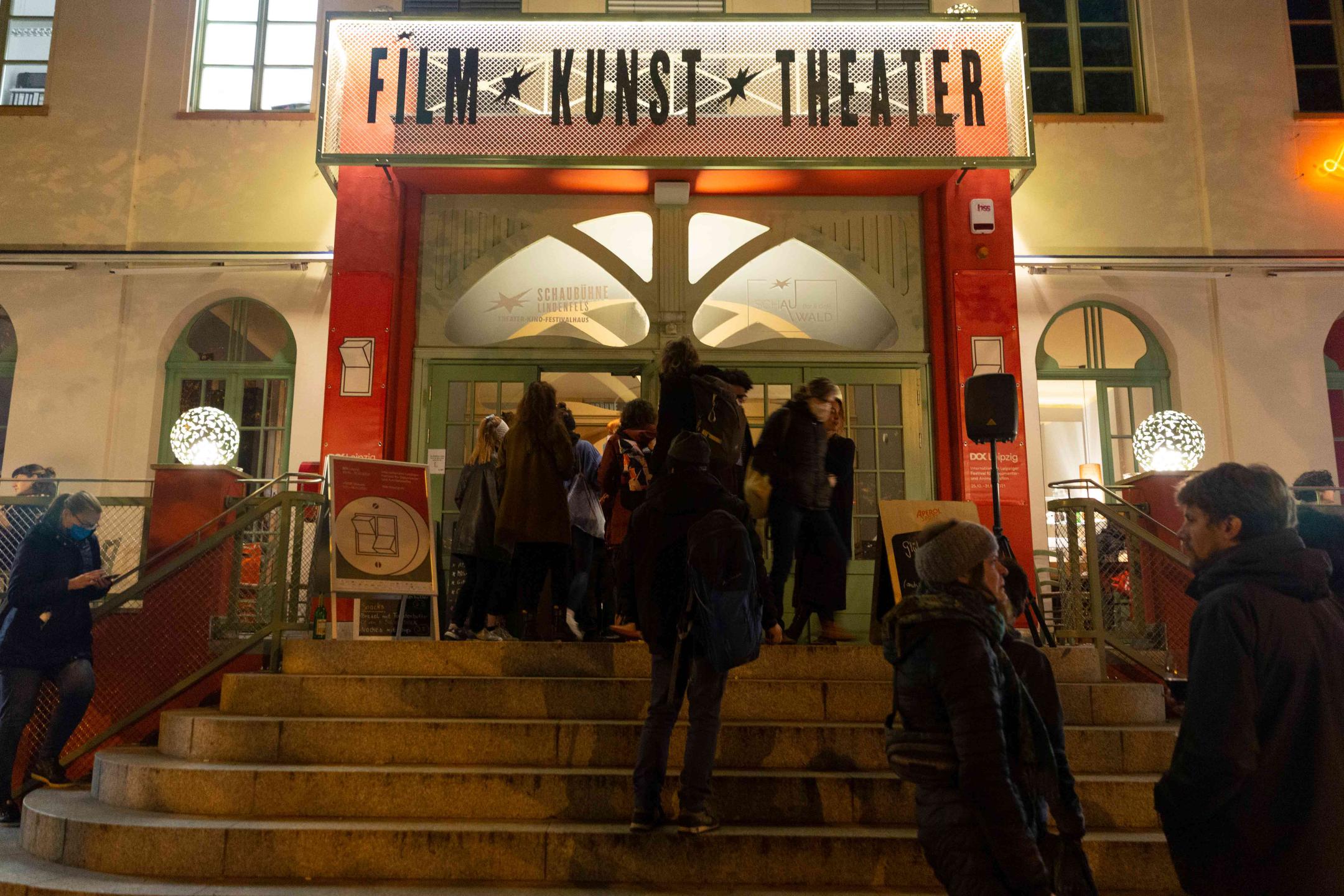Mehrere Personen stehen Schlange an einer Treppe, die zu einem großen beleuchteten Eingang führt. Darüber steht "Film * Kunst * Theater".