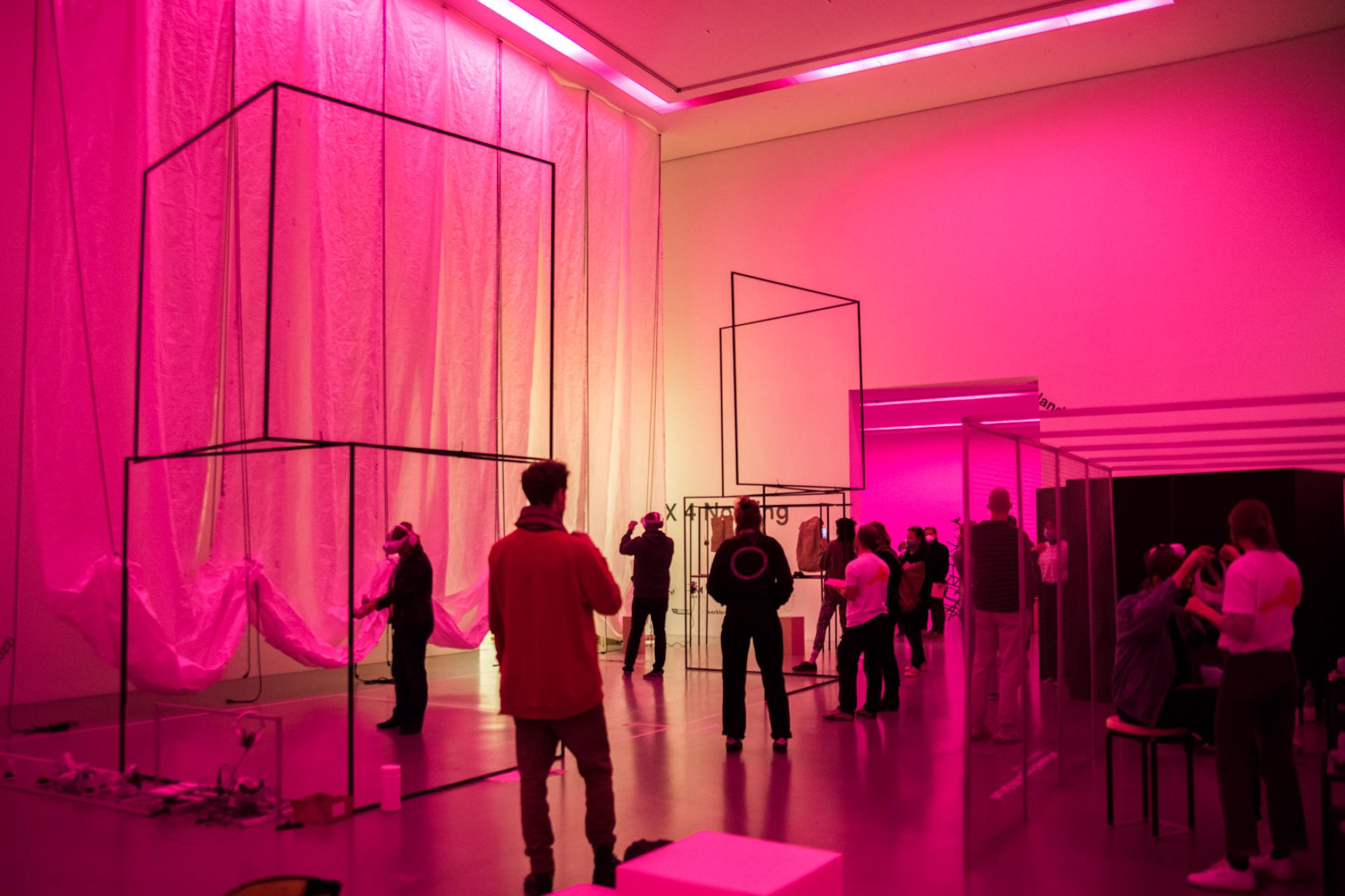 In einem Ausstellungsraum mit hohen Decken stehen und bewegen sich mehrere Menschen. Der Raum ist in pinkes Licht getaucht. Von der Decke hängt an einer Seite des Raums weiße, bauschige Stoffbahnen. Im Rahm stehen mehrere durchlässige Aufbauten aus Stahlgestängen.