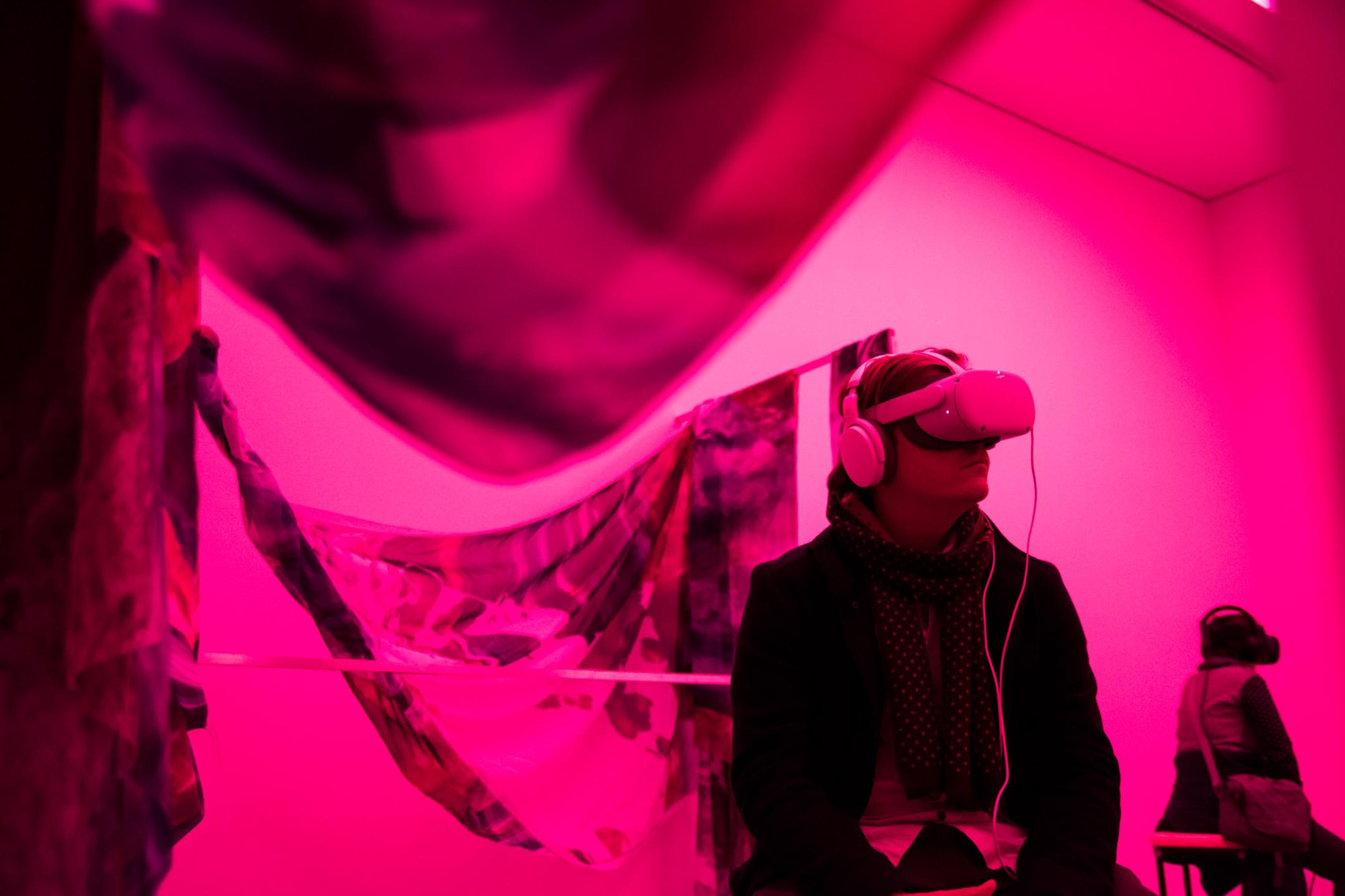 Ein Mann sitzt VR-Brille und Kopfhörern in einem Raum, der in pinkes Licht getaucht ist. Um ihn herum sind große Tücher mit schwarz-weißem Muster gespannt.