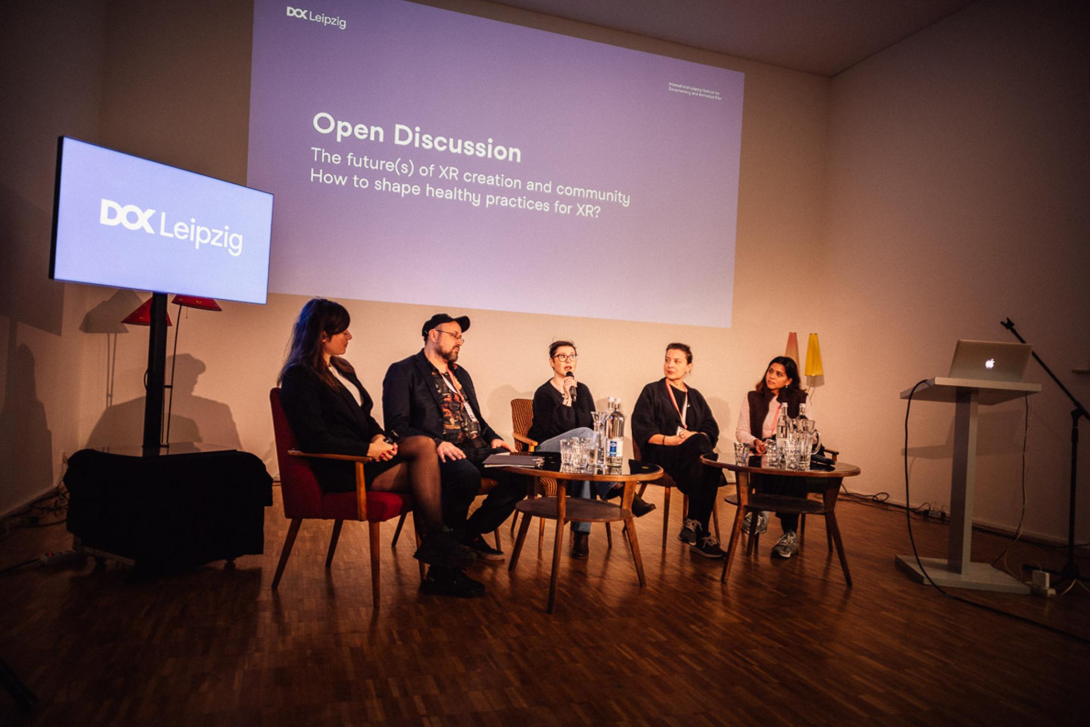 Five persons are sitting on sofa chairs on a discussion panel. The woman in the middle is talking into a microphone. In the background a blue info graphic is projected on the wall. It says: "Open discussion. The future(s) of XR creation and community."