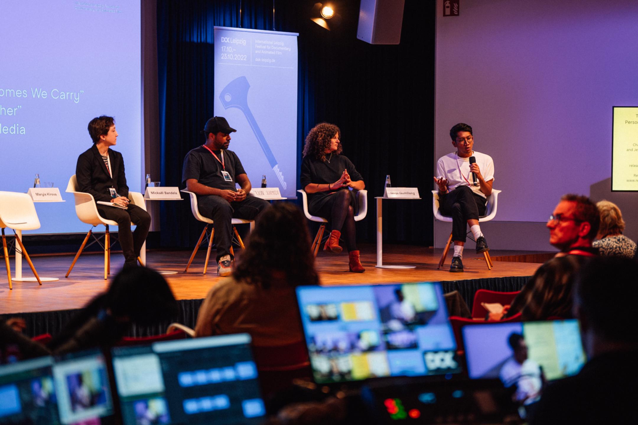 Four persons (two women, two men) are sitting on a panel. The man at the right is talking into a handheld microphone, the others are listening. In the front, the monitors of a live-editing suite light up in the dark.