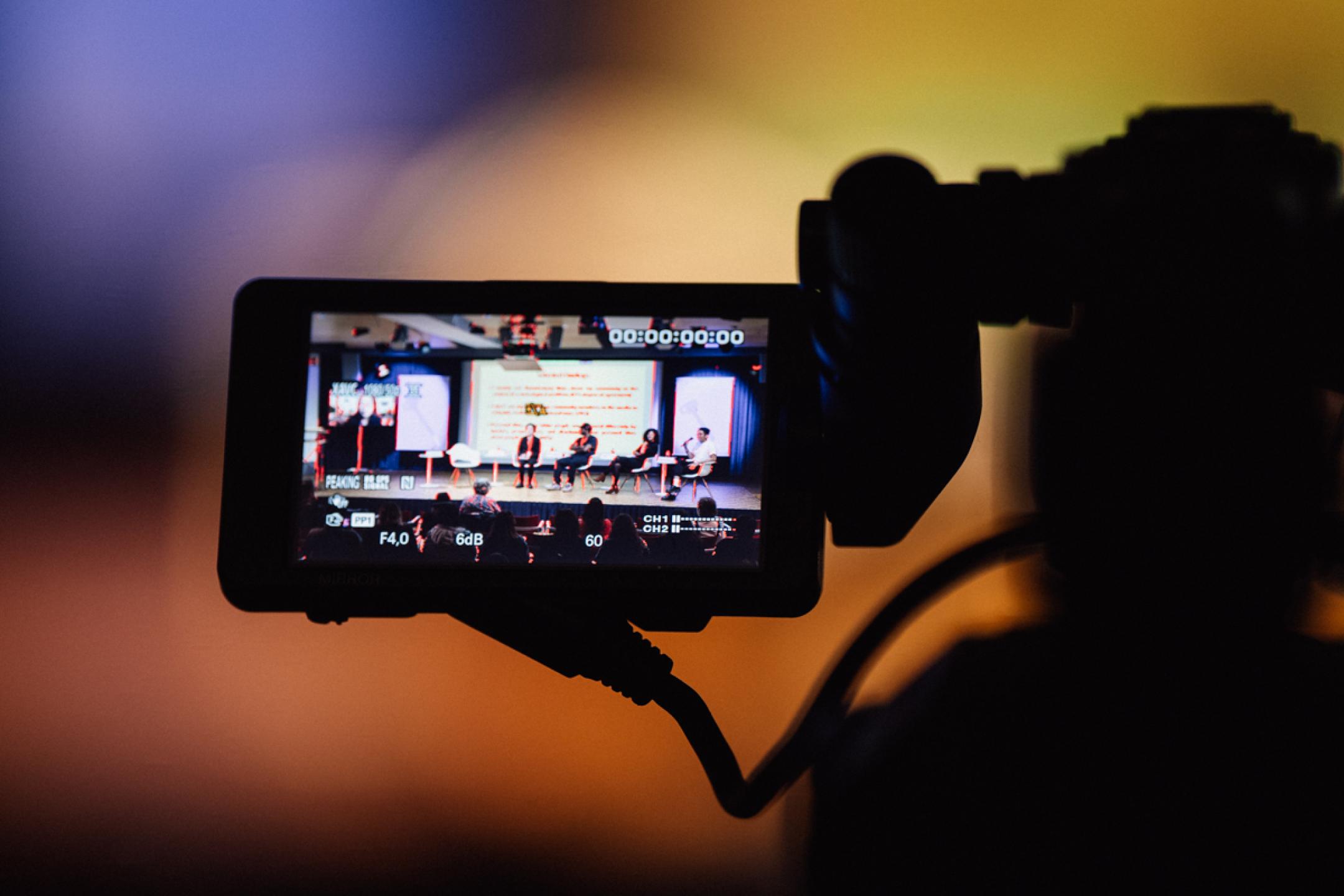 The monitor of a TV camera. On the screen the setting of a panel with four persons sitting on stage, one chair (left) is empty.