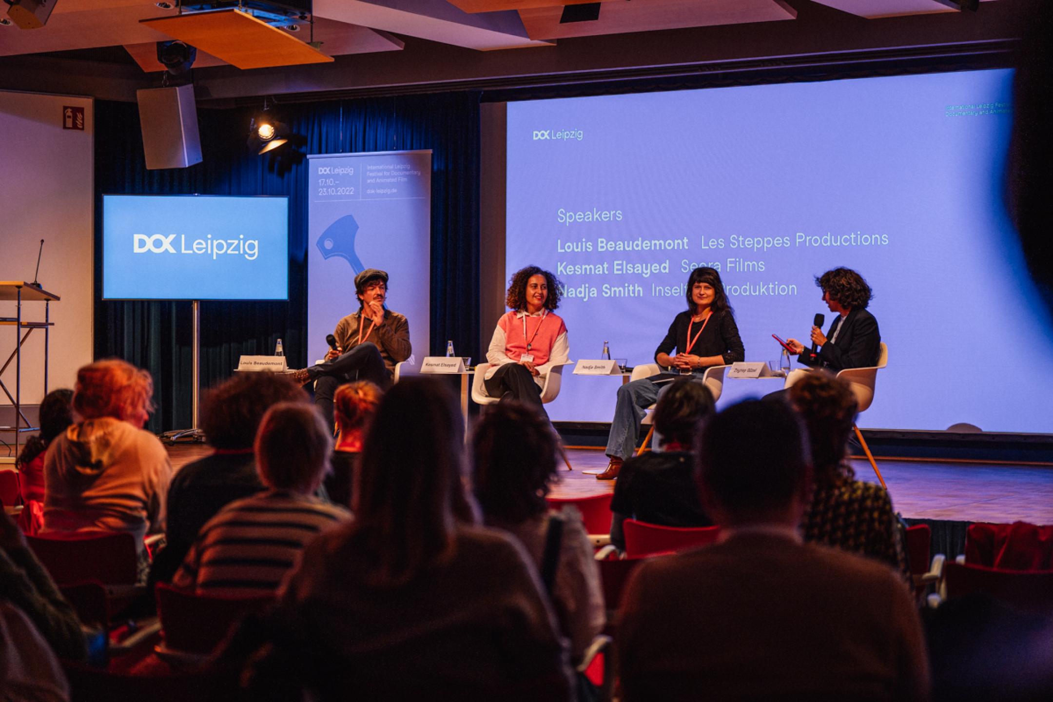 A talk panel with four persons sitting in chairs on stage: One man and three women.