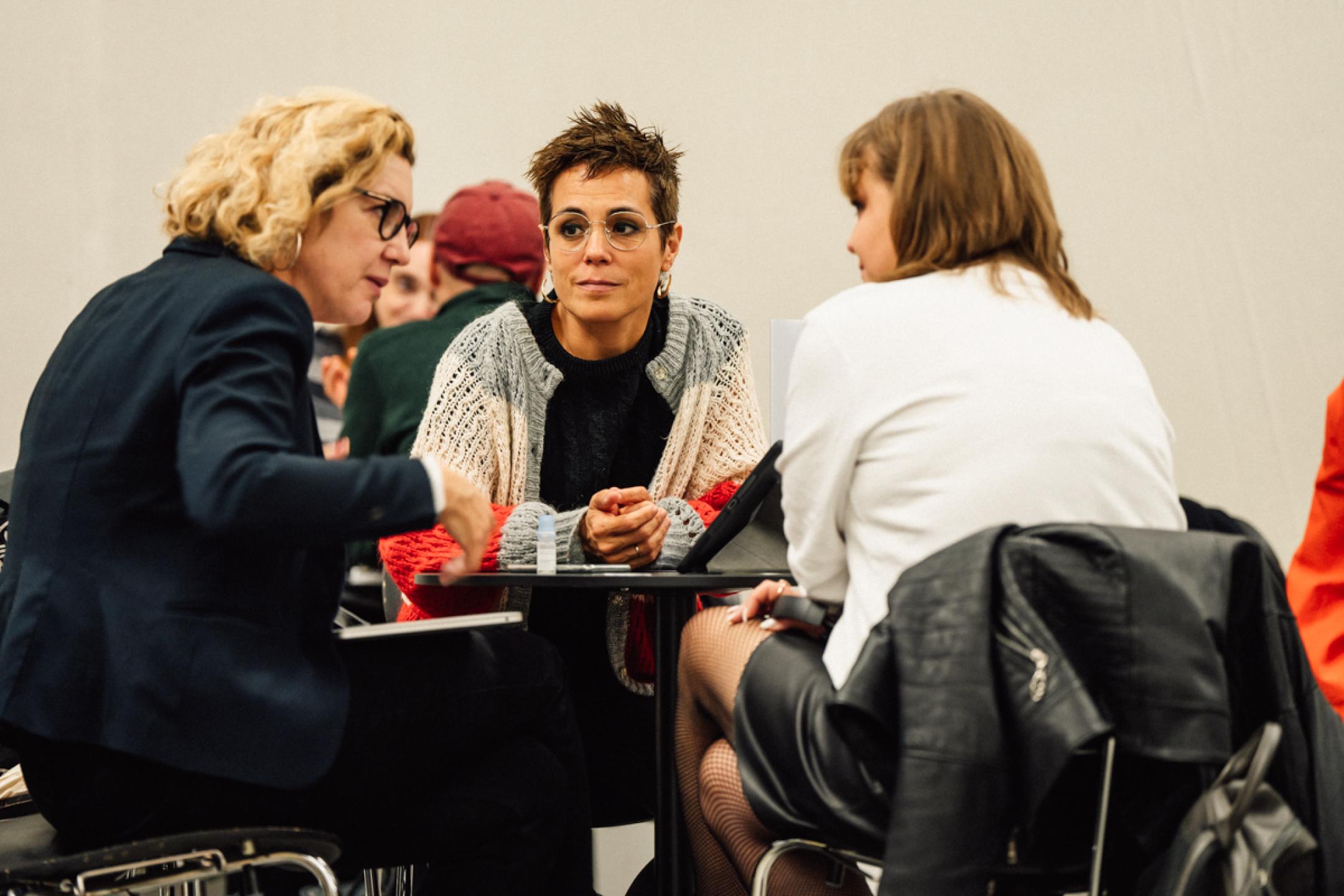Three women sit around a small round coffee table. The women at the left is speaking, the other two are listening.
