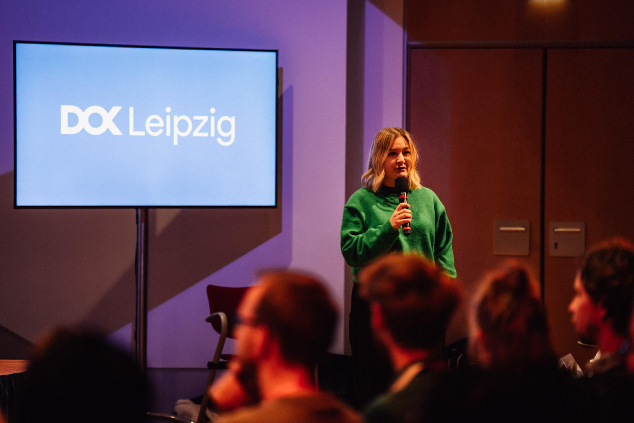 Eine Frau in einem grünen Pullover steht mit Handmikrofon vor einem blauen Bildschirm mit DOK Leipzig Logo.