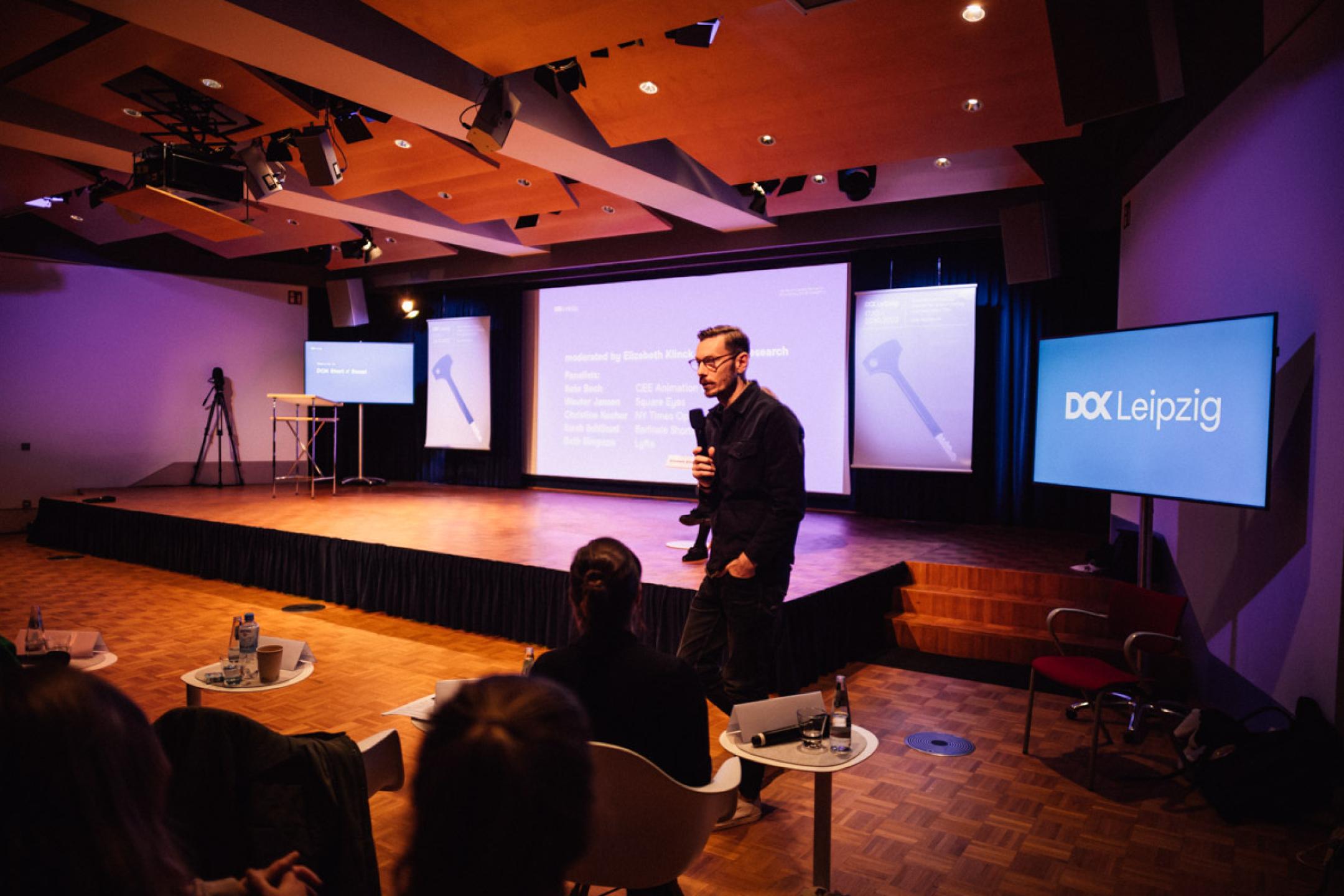 A man stands in front of an audience. In his back there is a huge screen in blue and on each side two big blue DOK Leipzig banners. The man speaks into a microphone.