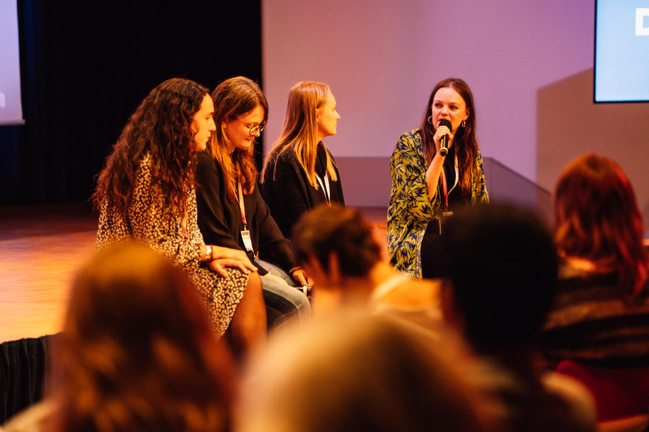 Moderator Brigid O’Shea sits in front of the stage and interviews three women who are sitting next to her.