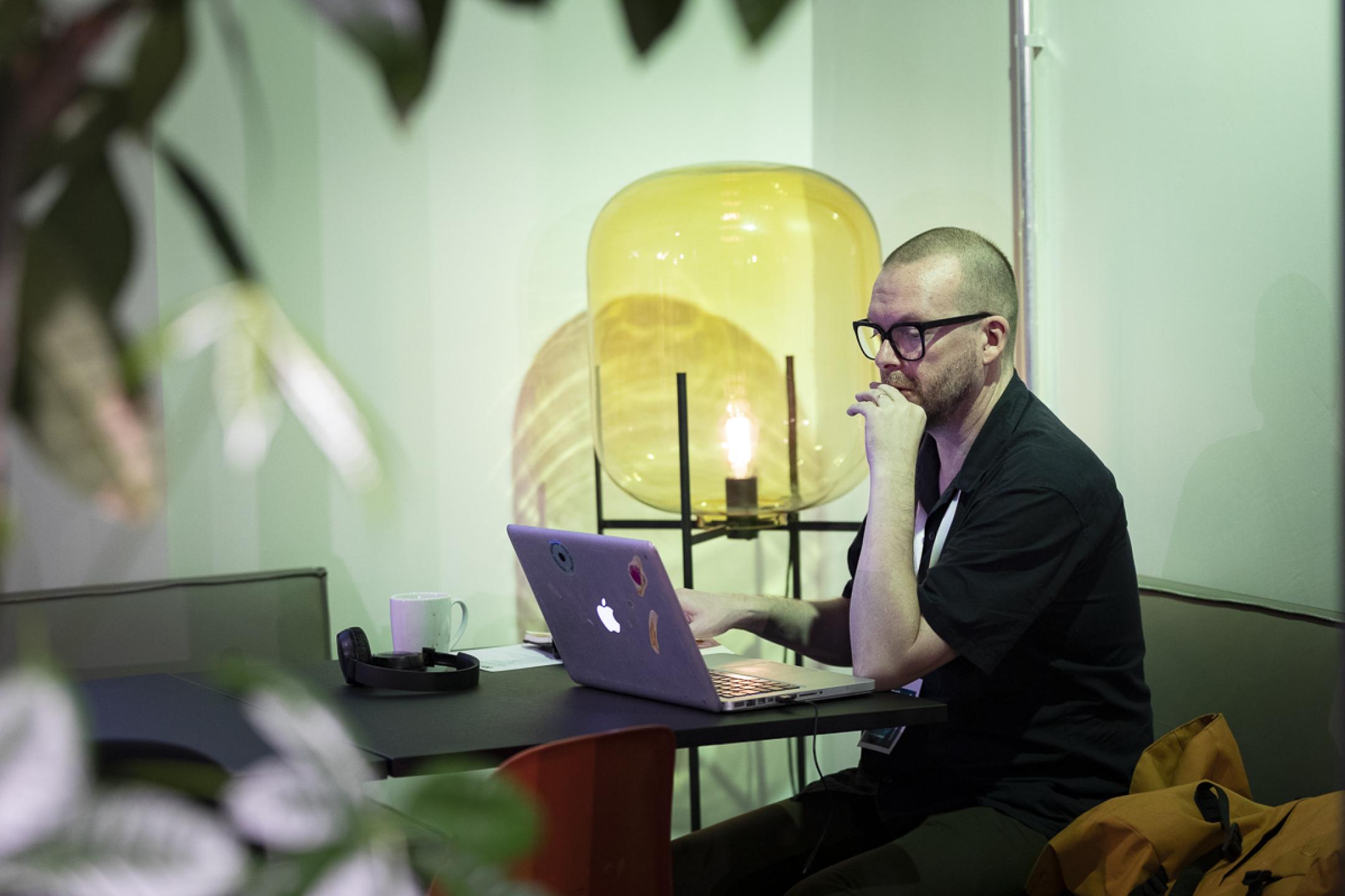 A man sits at a bistro table working on his laptop. In the background there is a huge lamp made of yellow glass.