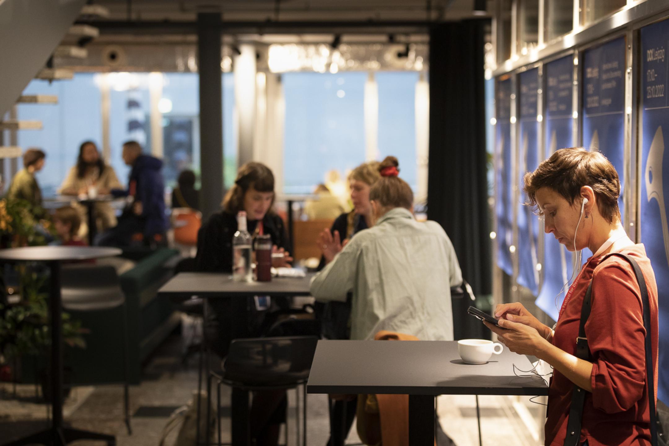 Eine Frau mit kurzen Haaren steht an einem Bistrotisch und blickt auf ihr Smartphone. Sie trägt Kopfhörer. Im Hintergrund sitzen mehrere Personen an weiteren Bistrotischen.