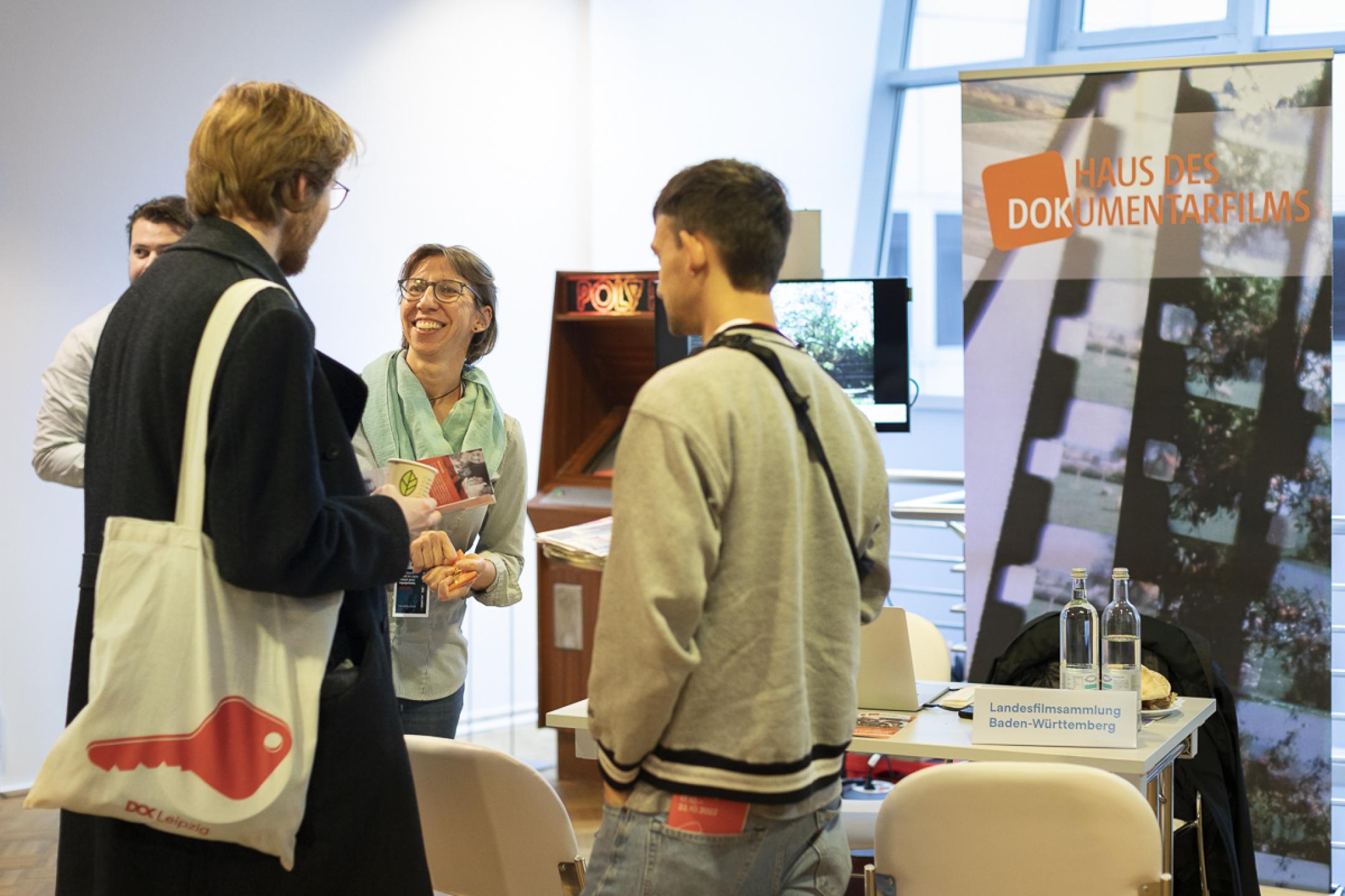 A woman is talking to two men. She stands next to a table with a screen behind and a poster of "Haus des Dokumentarfilms. 