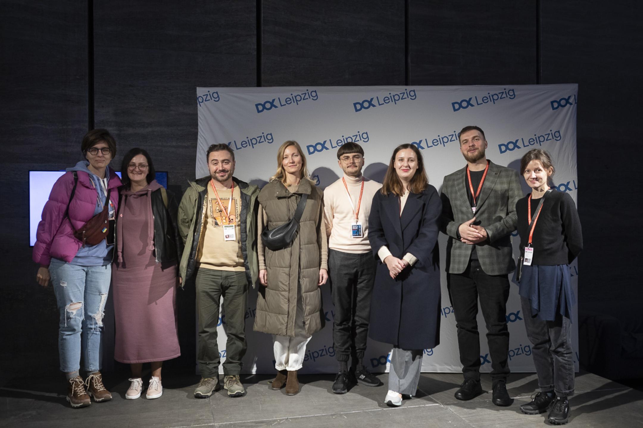 Eight persons (five women and three men) stand in front of a logo wall with the DOK Leipzig logo and pose for a group picture.