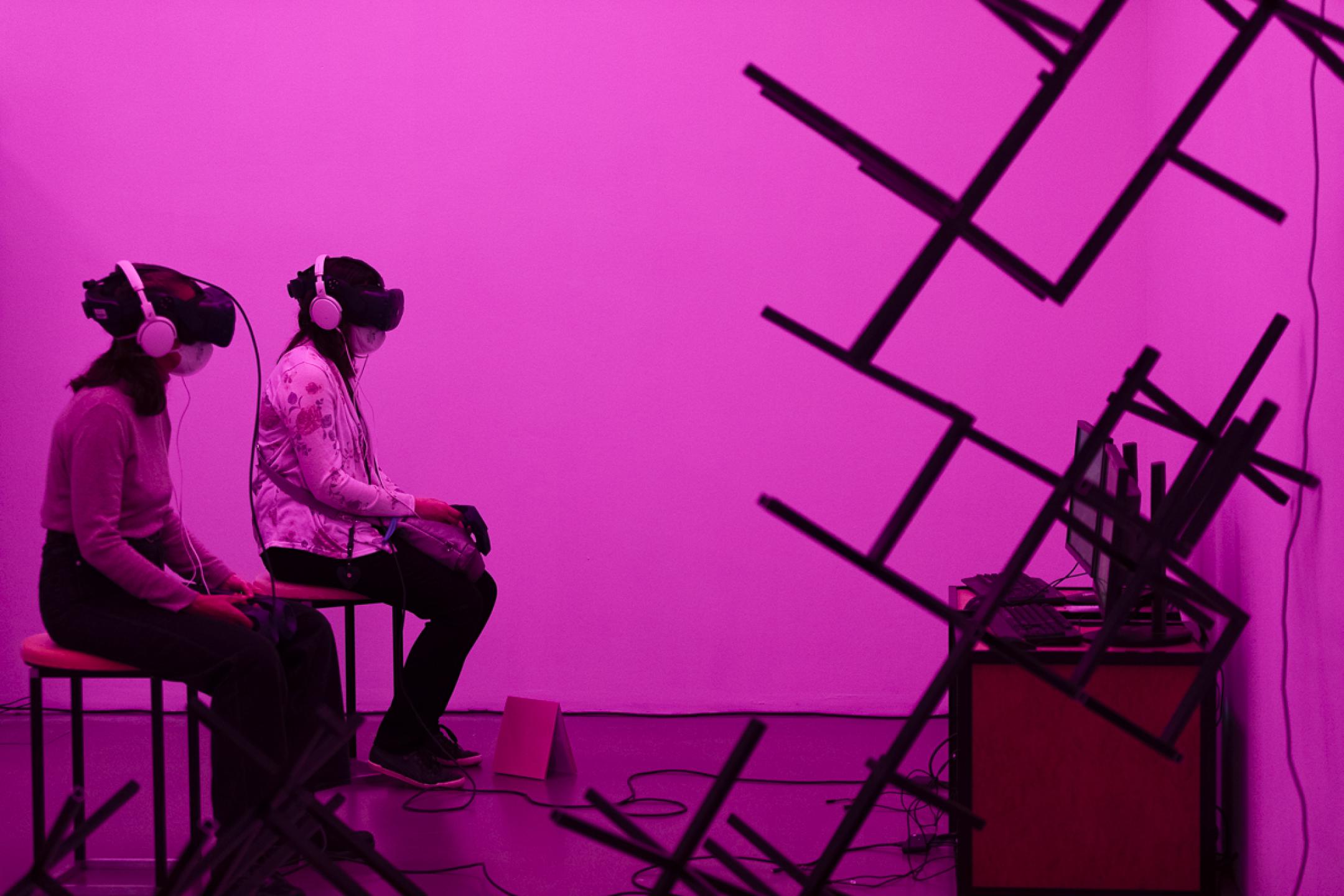 Zwei Frauen sitzen nebeneinander auf Hockern, beide tragen VR-Brillen. Der Raum um sie herum ist in pinkes Licht getaucht, an der Wand hängen abstrakte Gebilde aus Stahlgestängen.