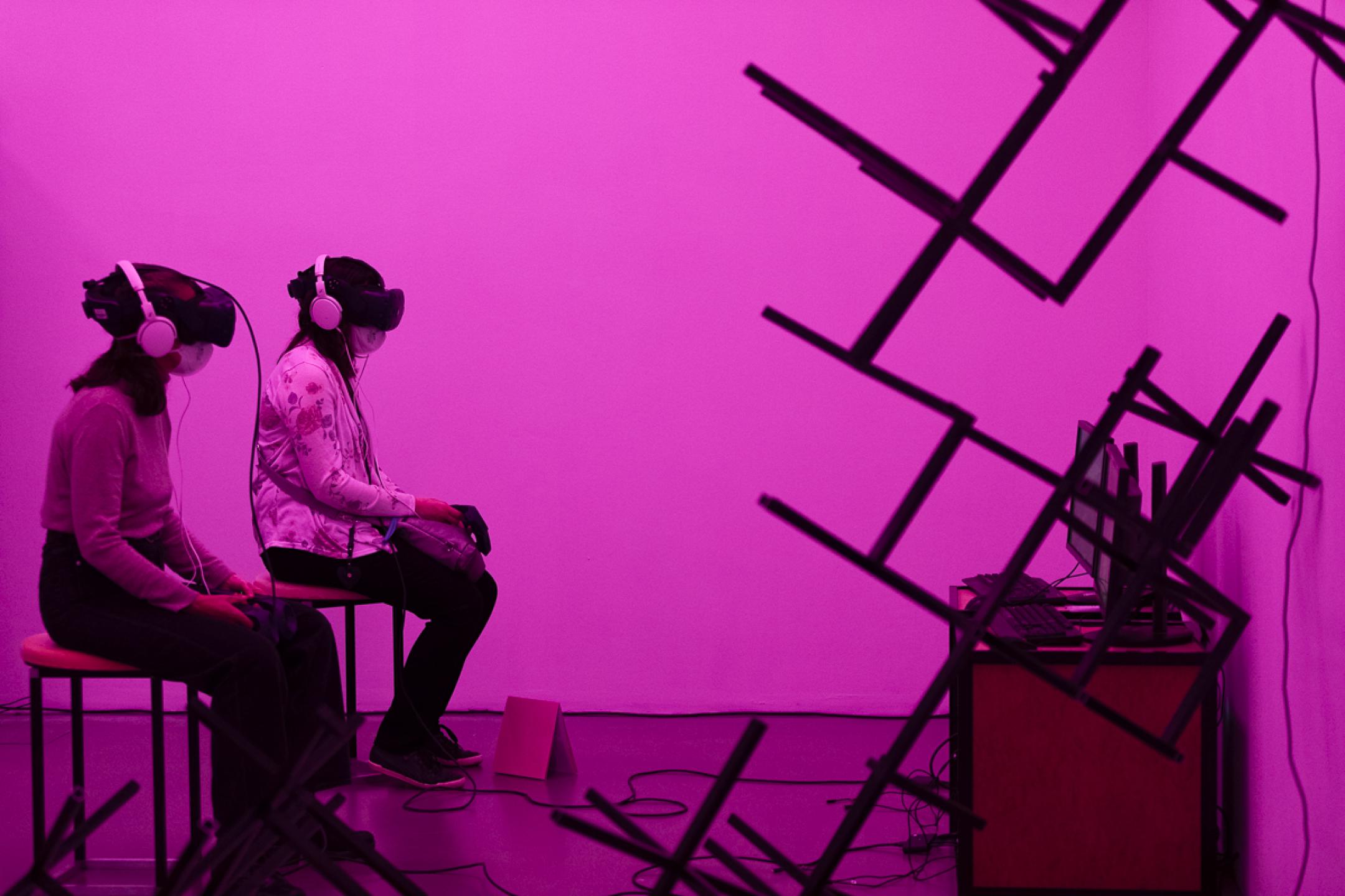 Zwei Frauen sitzen nebeneinander auf Hockern, beide tragen VR-Brillen. Der Raum um sie herum ist in pinkes Licht getaucht, an der Wand hängen abstrakte Gebilde aus Stahlgestängen.