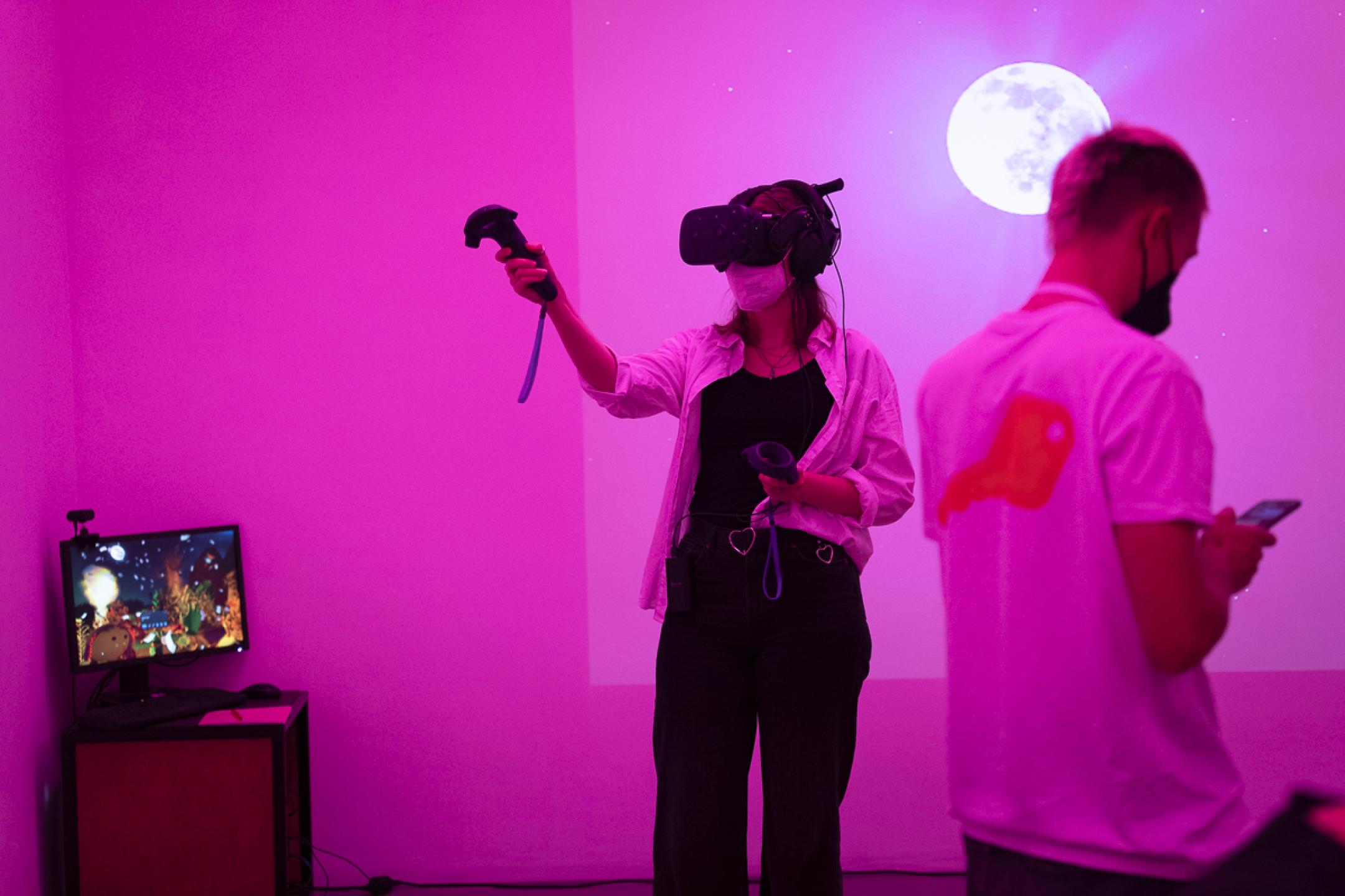 Eine Frau mit VR-Brille steht in einem pinken Raum und zeigt mit ihrer Hand mit einem VR-Controller nach oben. Hinter ihr wird ein leuchtender Planet an die Wand projiziert. In einer Ecke steht ein Bildschirm, der eine Landschaft mit Sternenhimmel bei Nacht zeigt.