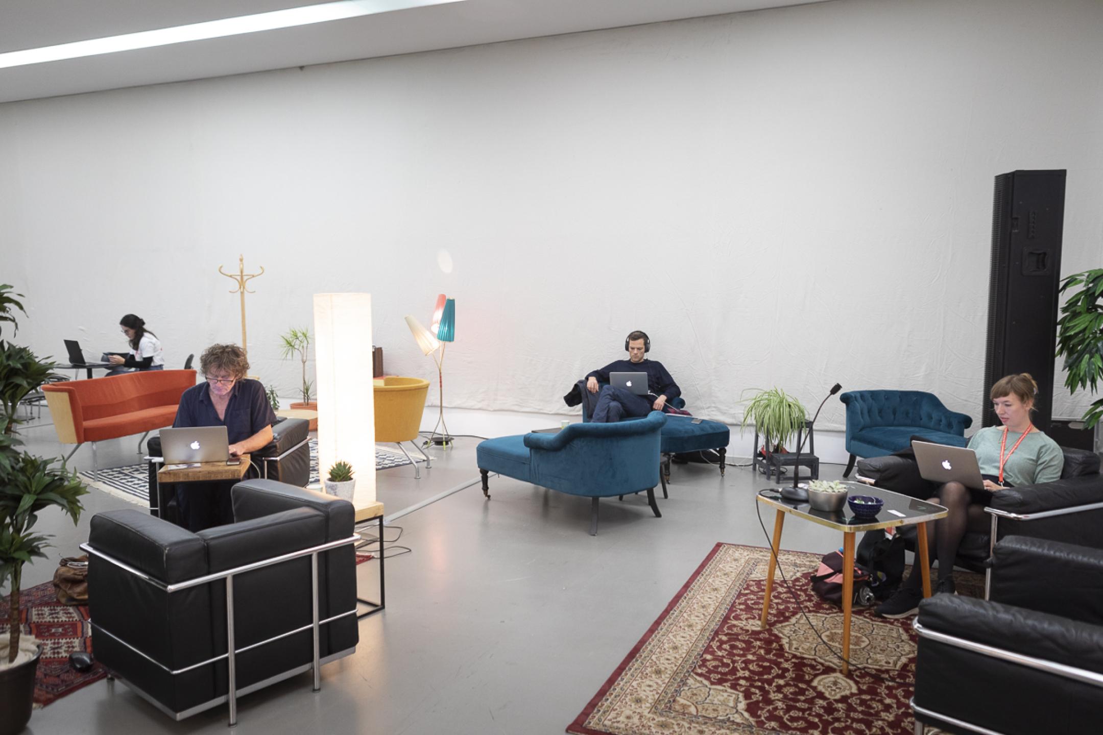 In a huge room, several sofa chairs are arranged around coffee tables. Three people sit with their laptops open, each for themselves at a table.