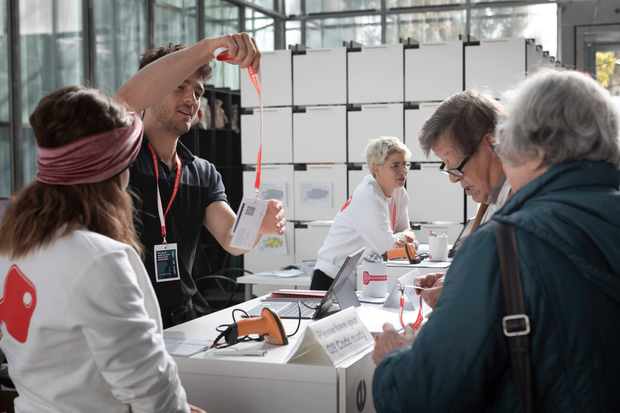 Mehrere Personen stehen um zwei weiße Tresen. Ein Mann hält ein Schlüsselband mit einem bedruckten Akkreditiertenausweis in die Höhe.
