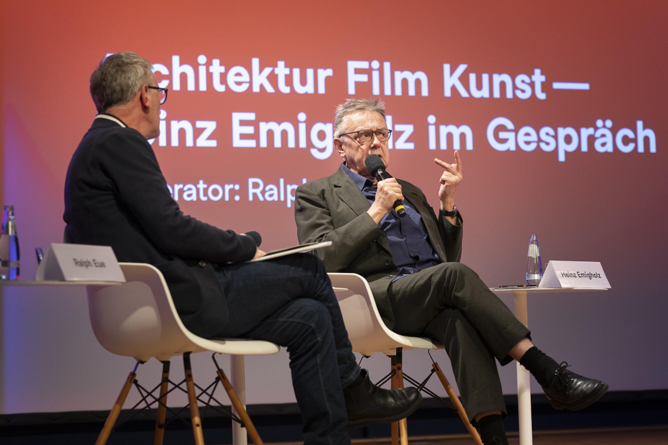 Filmmaker Heinz Emigholz sits next to another man on a panel. He leans back in his chair and speaks into a handheld microphone while gesturing with his other hand.