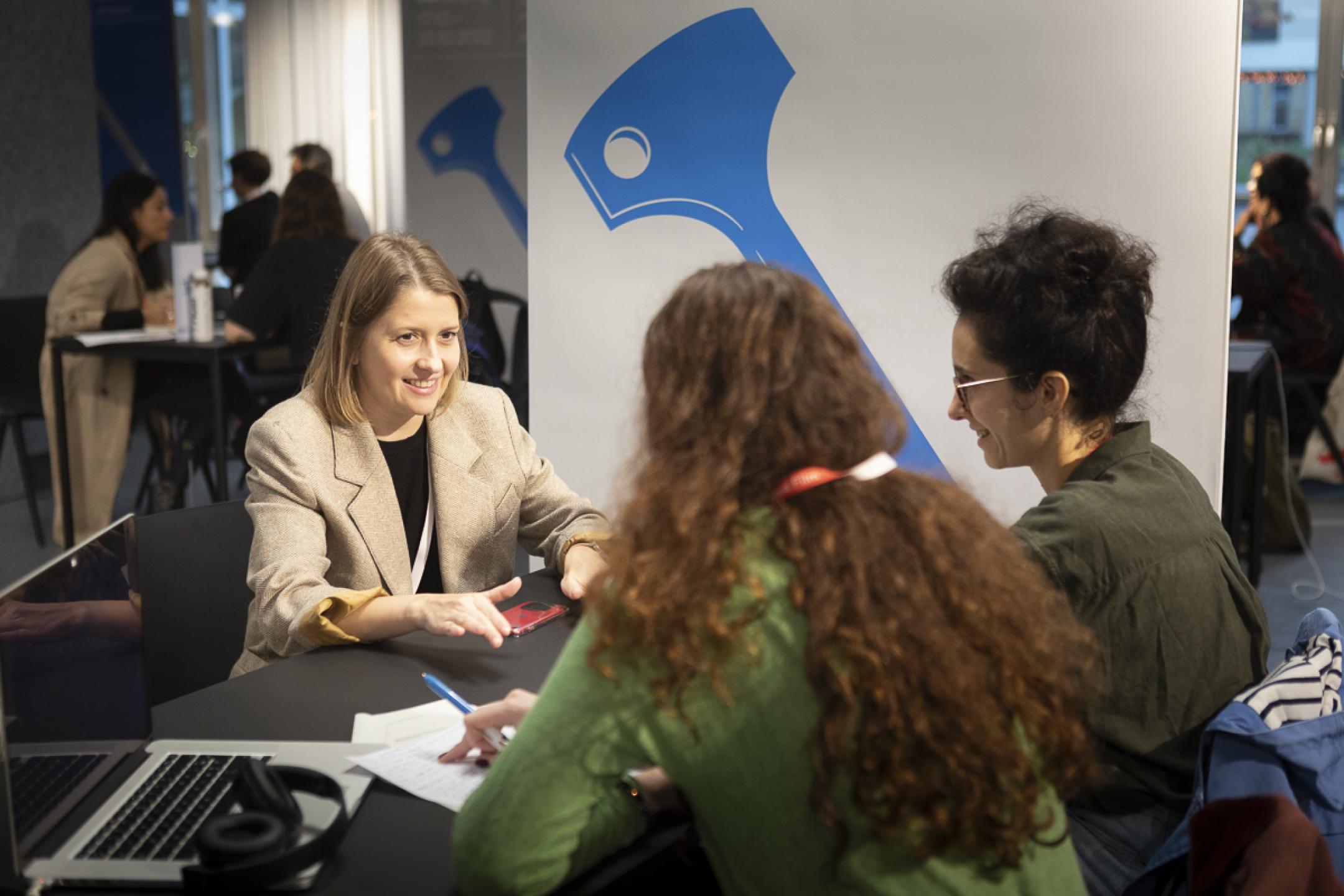 A woman wits opposite to two other women at a table. She is smiling brightly while gesturing with her hands. The other two listen attentively and take notes on a paper.