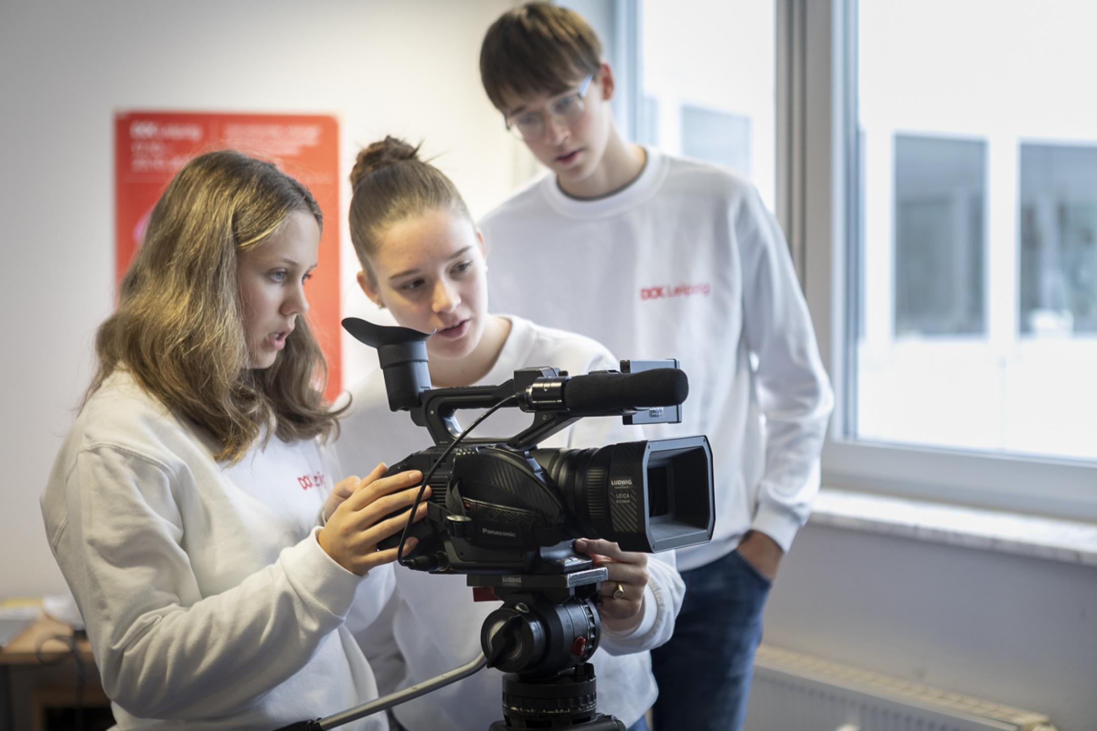 Drei Jugendliche – zwei Mädchen und ein Junge – beugen sich über eine Videokamera auf einem Stativ. 