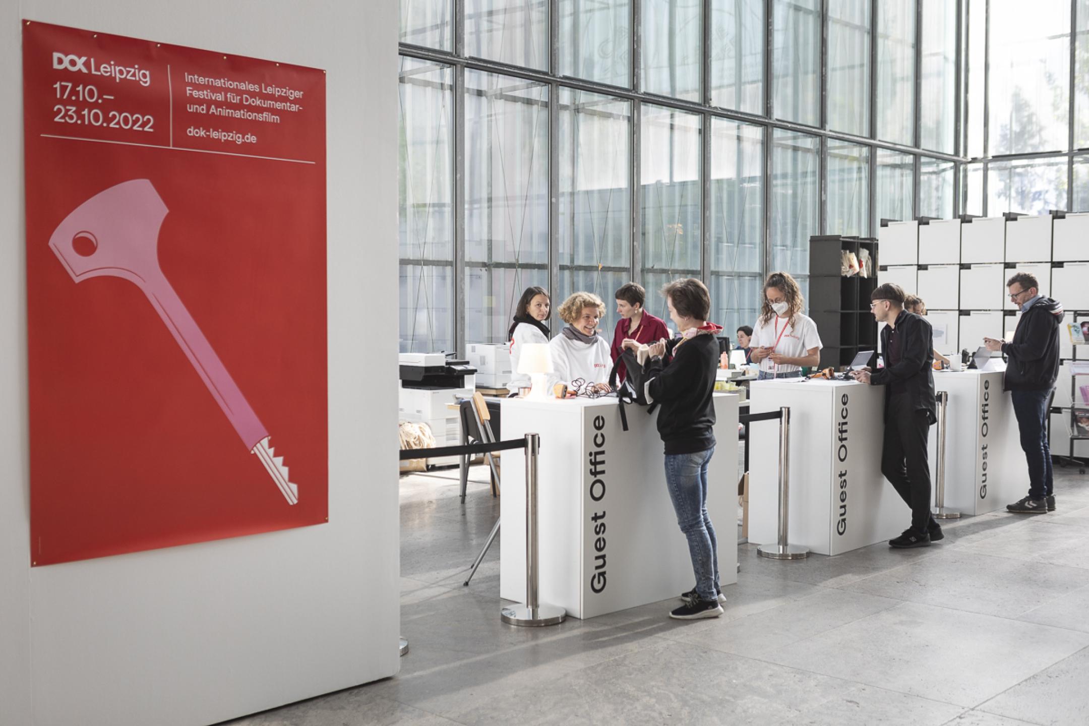 Three white reception counters at the festival centre: At each counter a team member is talking to a festival visitor. At the left the festival poster is hanging at a huge white wall.