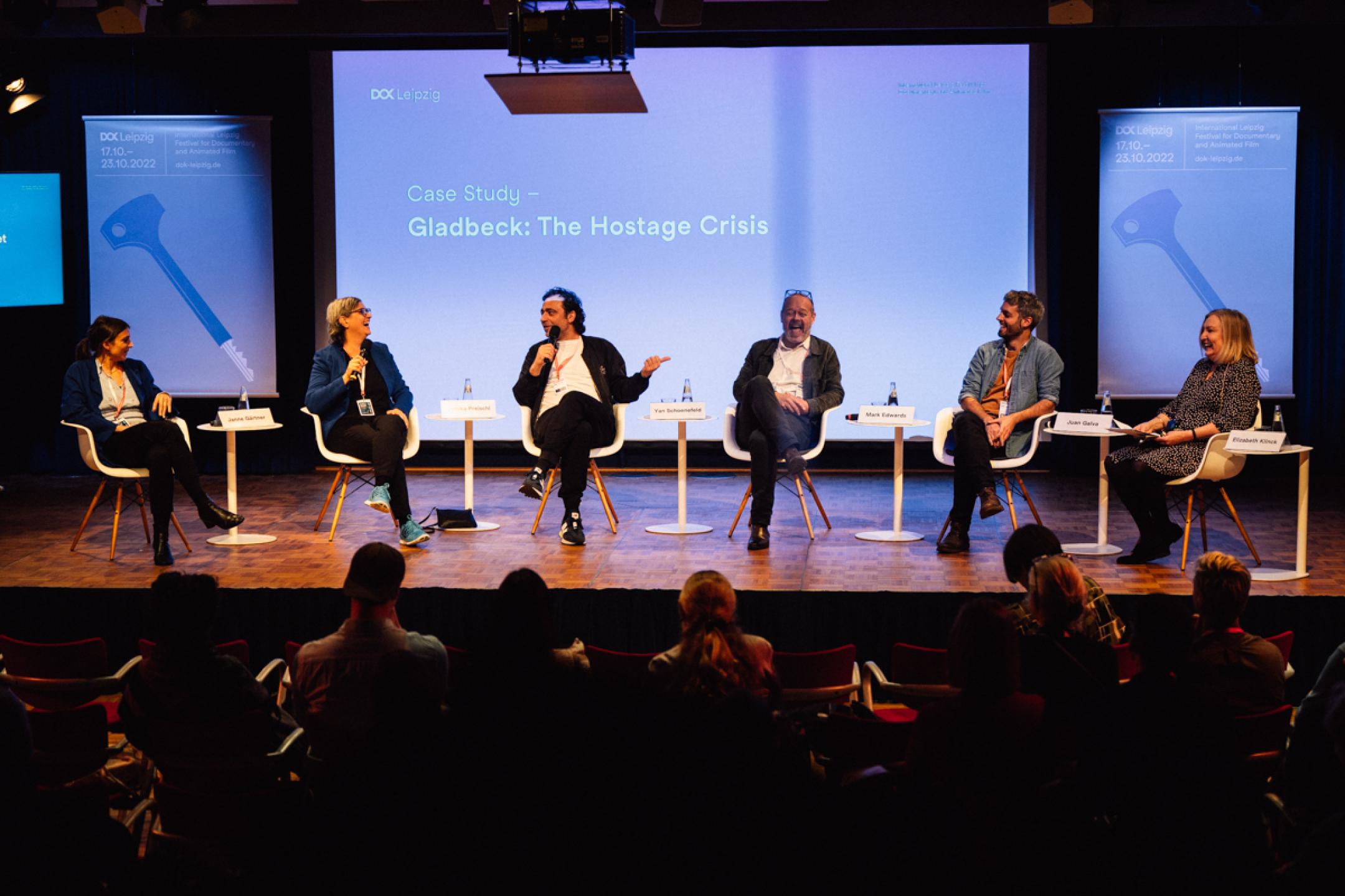 Six persons (two women, four men) sit on a panel in front of an audience. The six are all laughing out loud. In their background a huge screen with text: "Dok Industry. Gladbeck Case Study" 