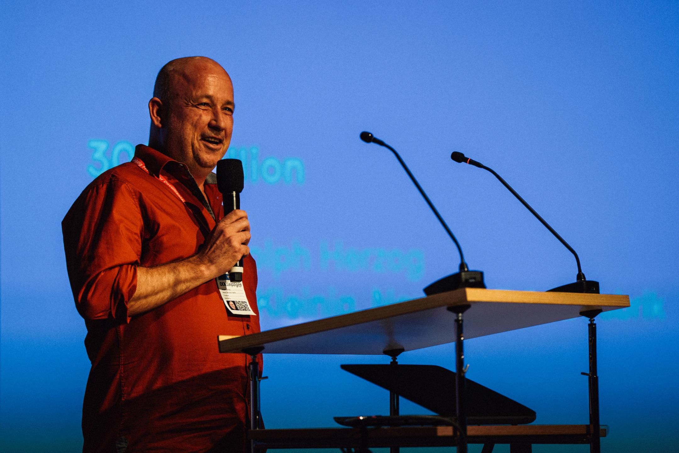 A men with a bald head and in an orange shirt stands at a speech desk and speaks into a microphone that he is holding in his hand.