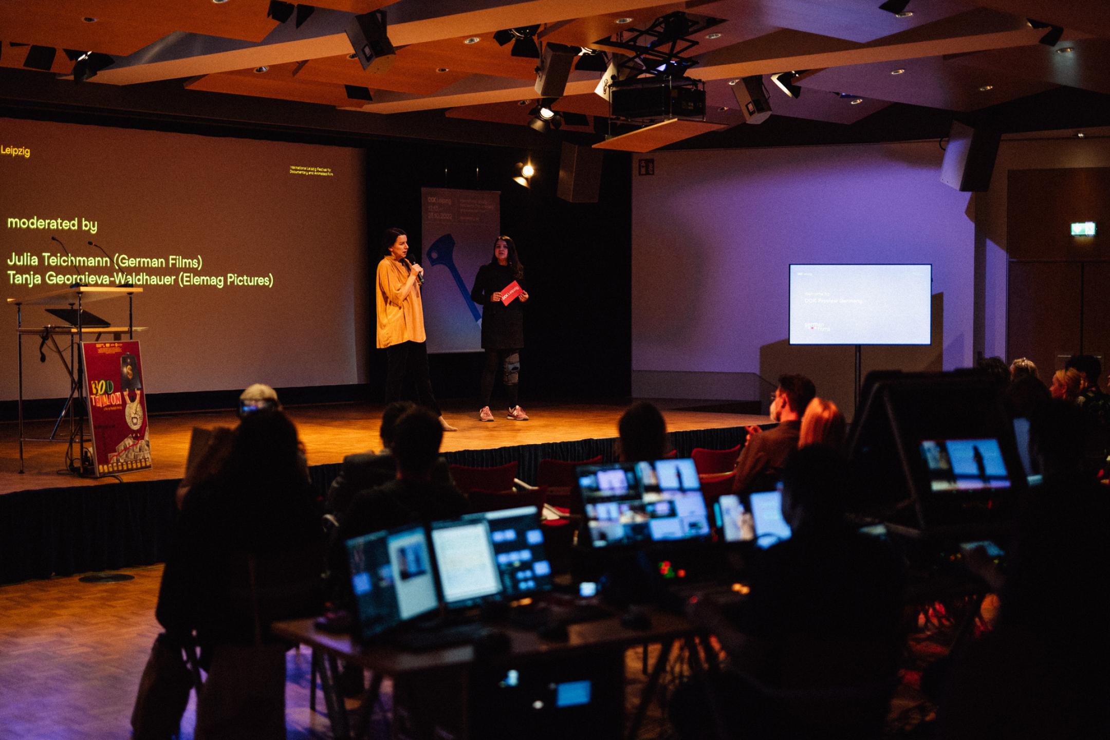 Two women are presenting on a stage in a conference room. In the middle of the audience is a live editing suite with several monitors. The monitors light up. 