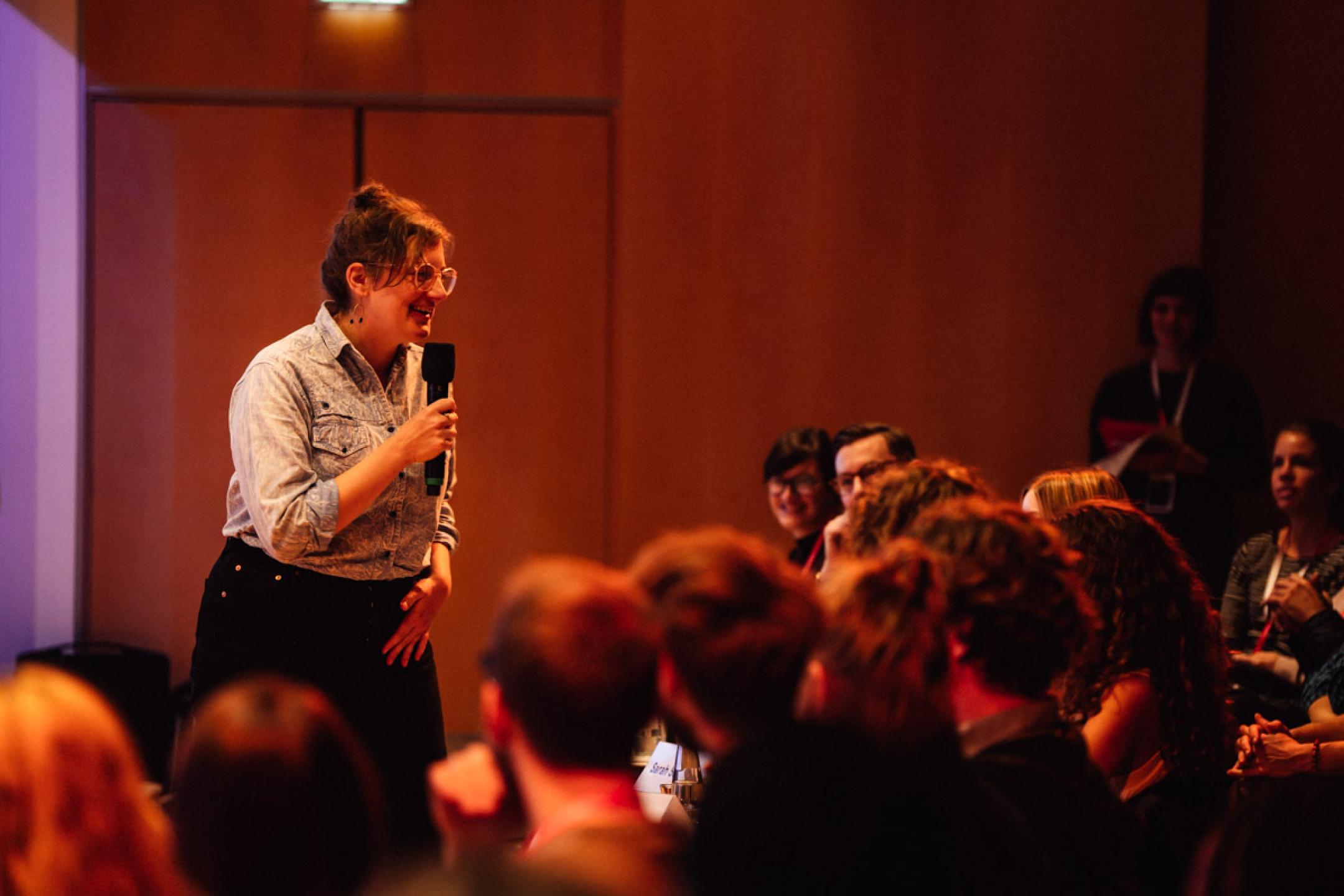 A woman in a jeans shirt stands in front of an audience. She holds a microphone in her hands and is laughing.