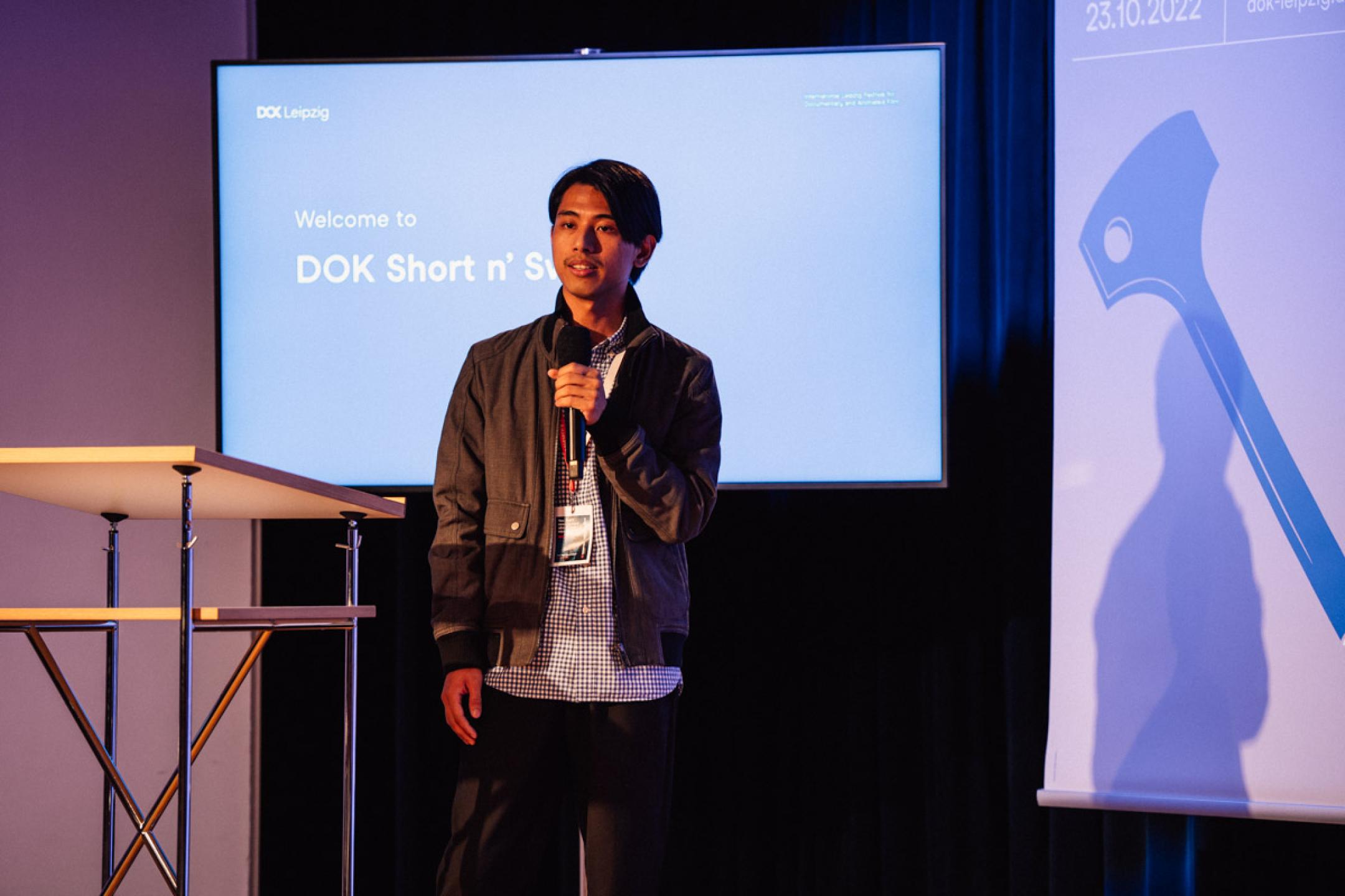 A young man with a black jacket and a Dok Leipzig badge stands on stage with a microphone. In his back a huge screen with text: "Welcome to Dok Short and Sweet."